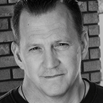 Black and white portrait of a man with short, slicked-back hair, standing in front of a brick wall.