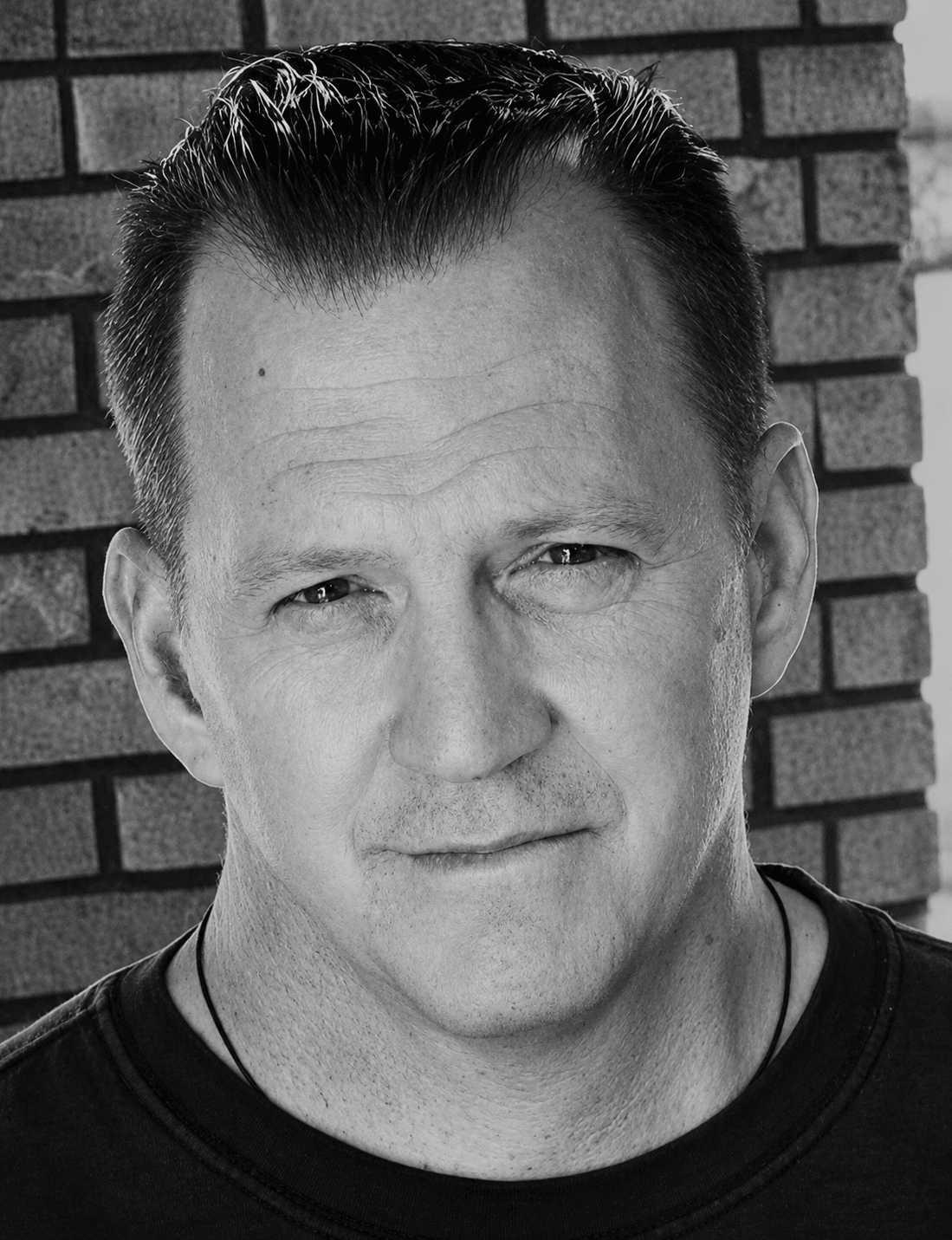 Black and white portrait of a man with short, slicked-back hair, standing in front of a brick wall.
