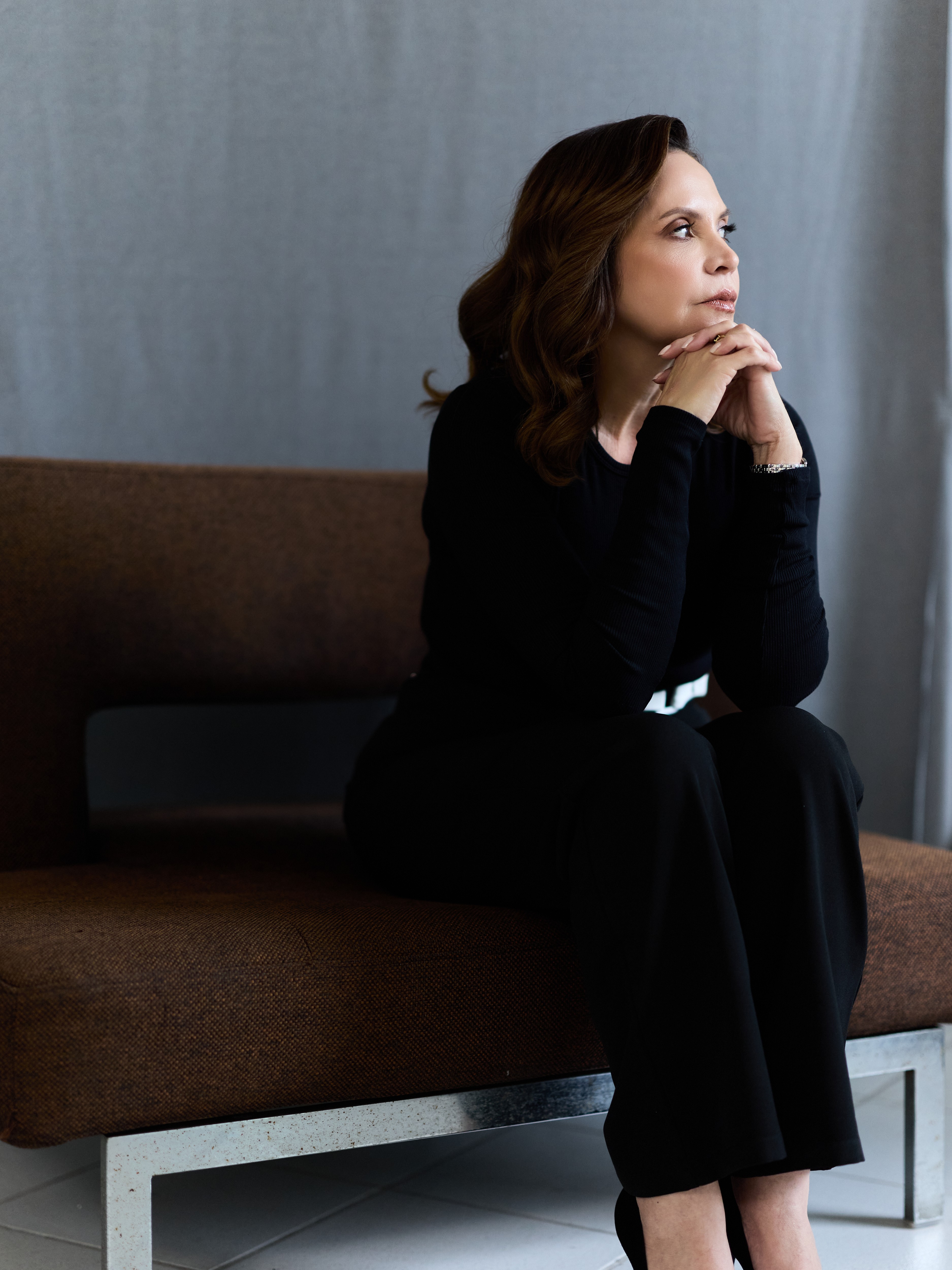 Woman with shoulder-length brown hair sitting on a brown sofa, looking thoughtfully to the side, hands clasped near her chin.