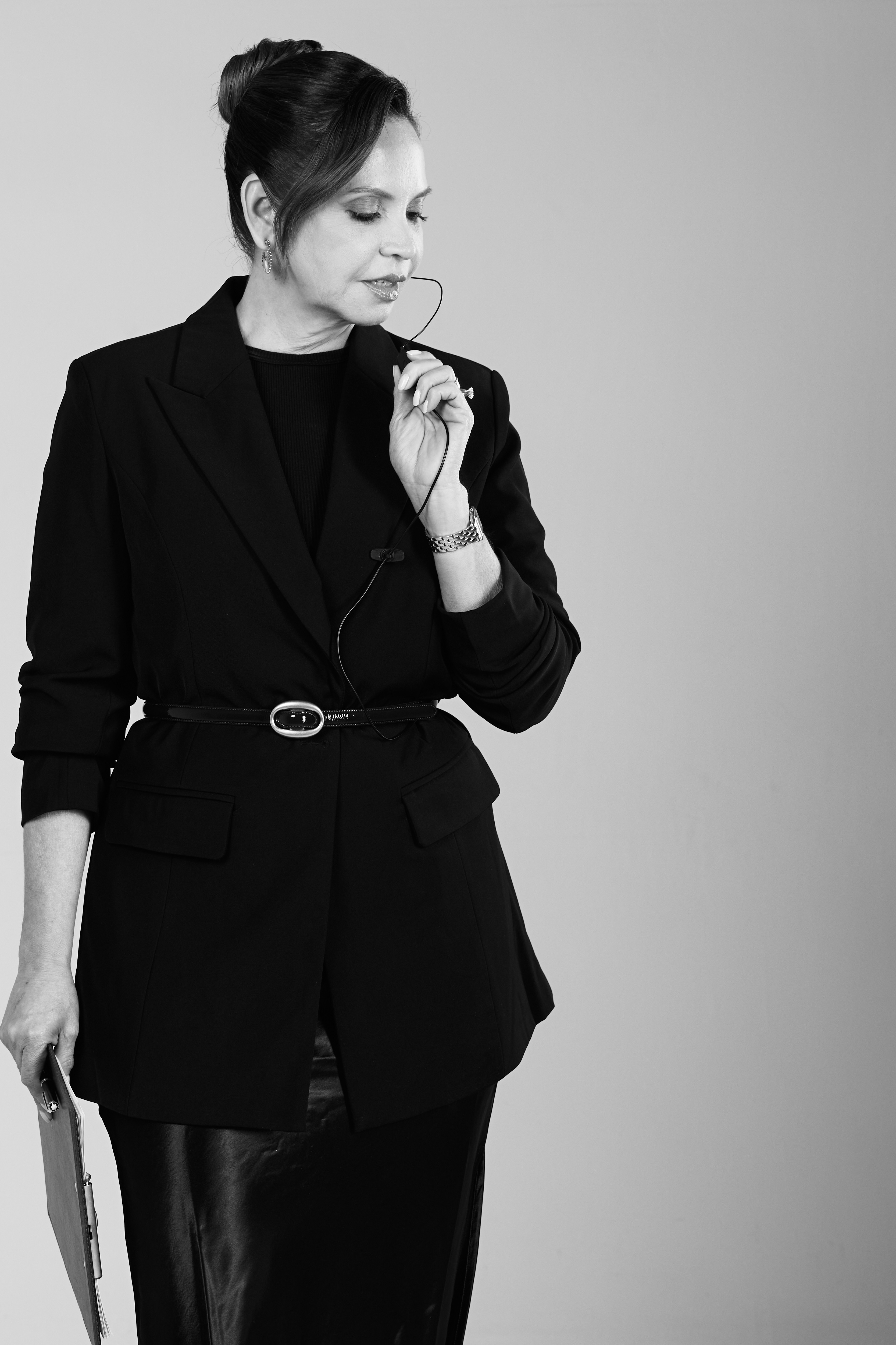 Woman in black blazer and skirt holding a book, looking down thoughtfully, against a plain background.
