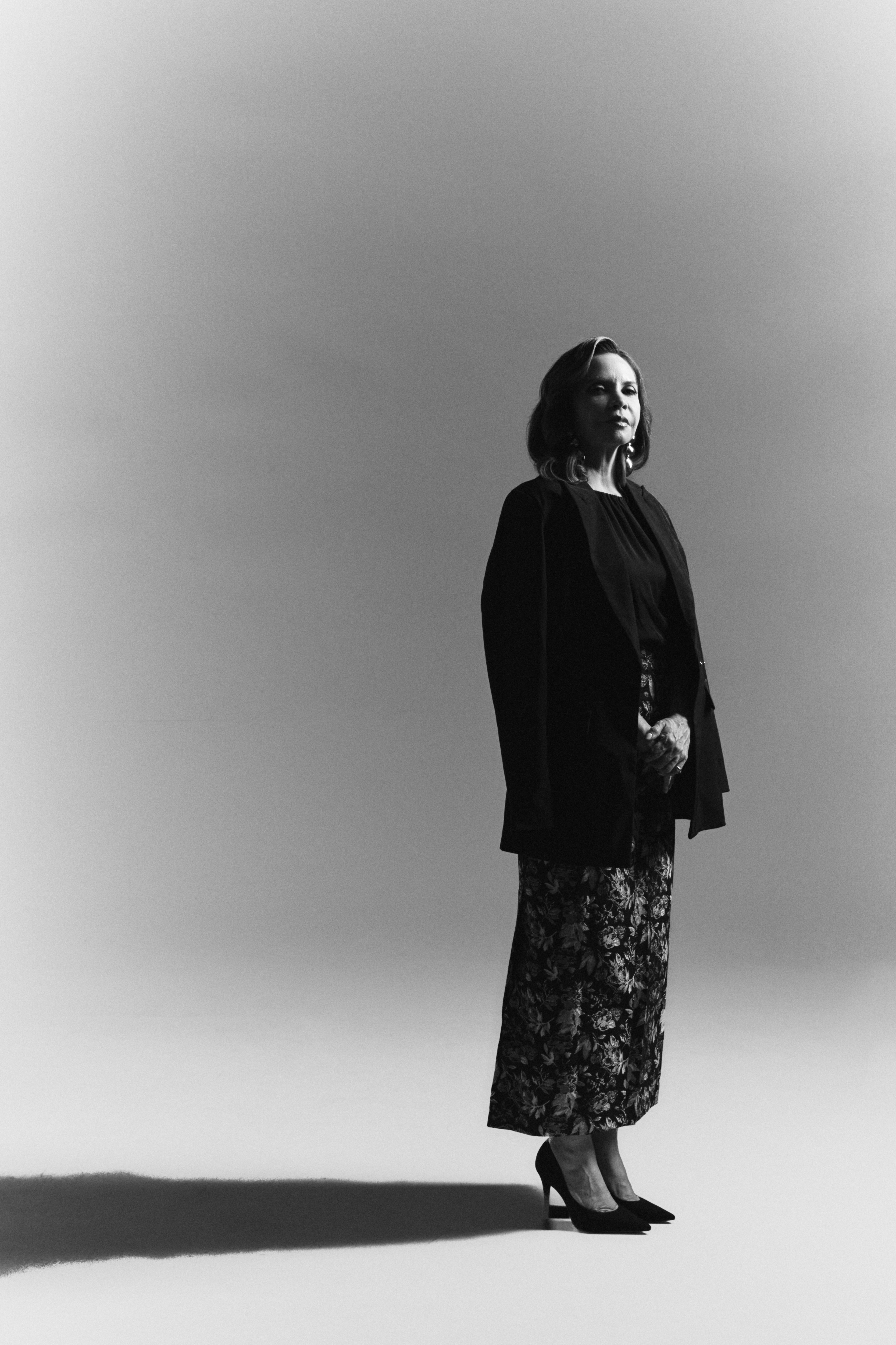 Woman standing outdoors with sky background, wearing a dark coat and patterned skirt, looking to the side.