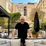 Man with glasses and black shirt leaning on outdoor furniture in a garden setting, with buildings in the background.