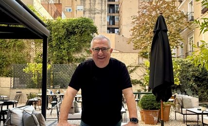 Man with glasses and black shirt leaning on outdoor furniture in a garden setting, with buildings in the background.