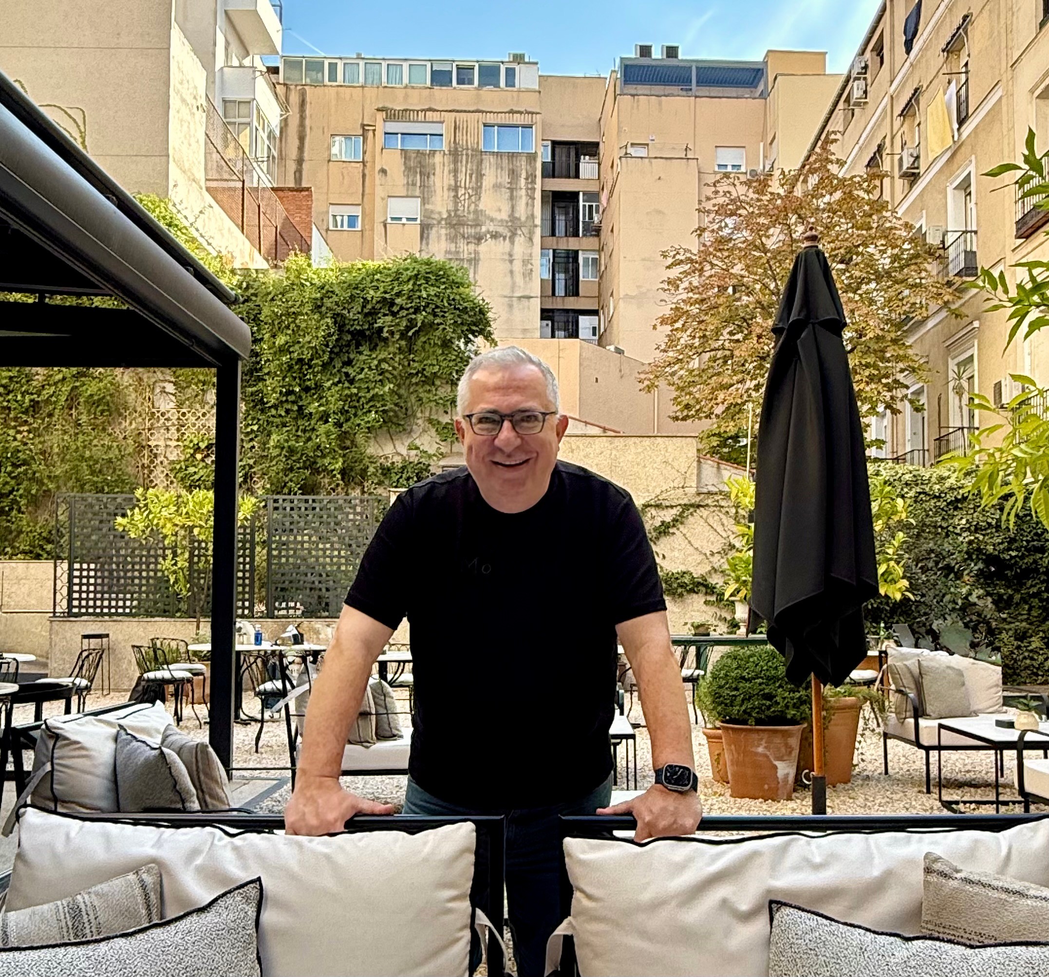 Man with glasses and black shirt leaning on outdoor furniture in a garden setting, with buildings in the background.