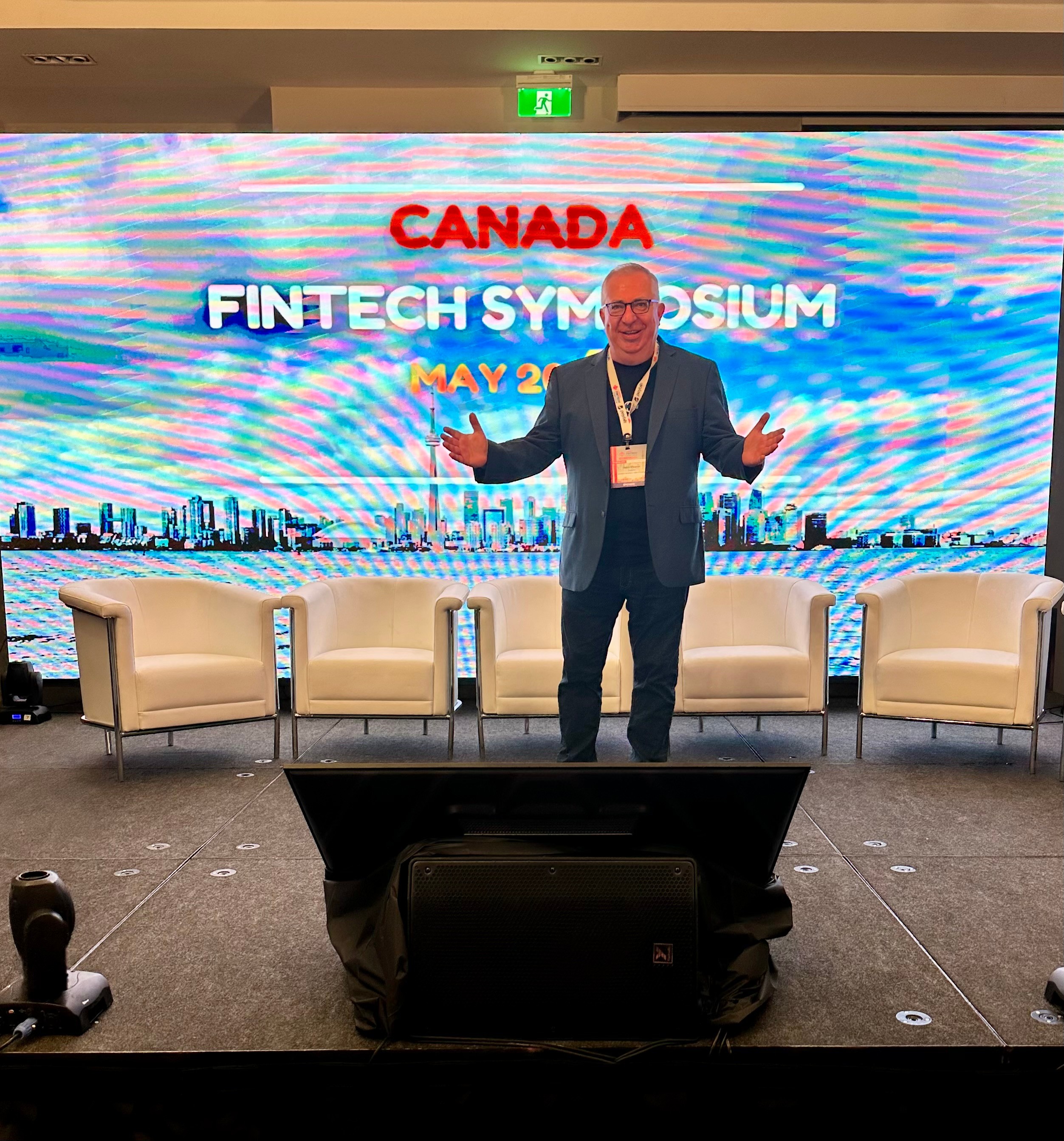 Man standing on stage with four chairs and a large screen behind him displaying event information.