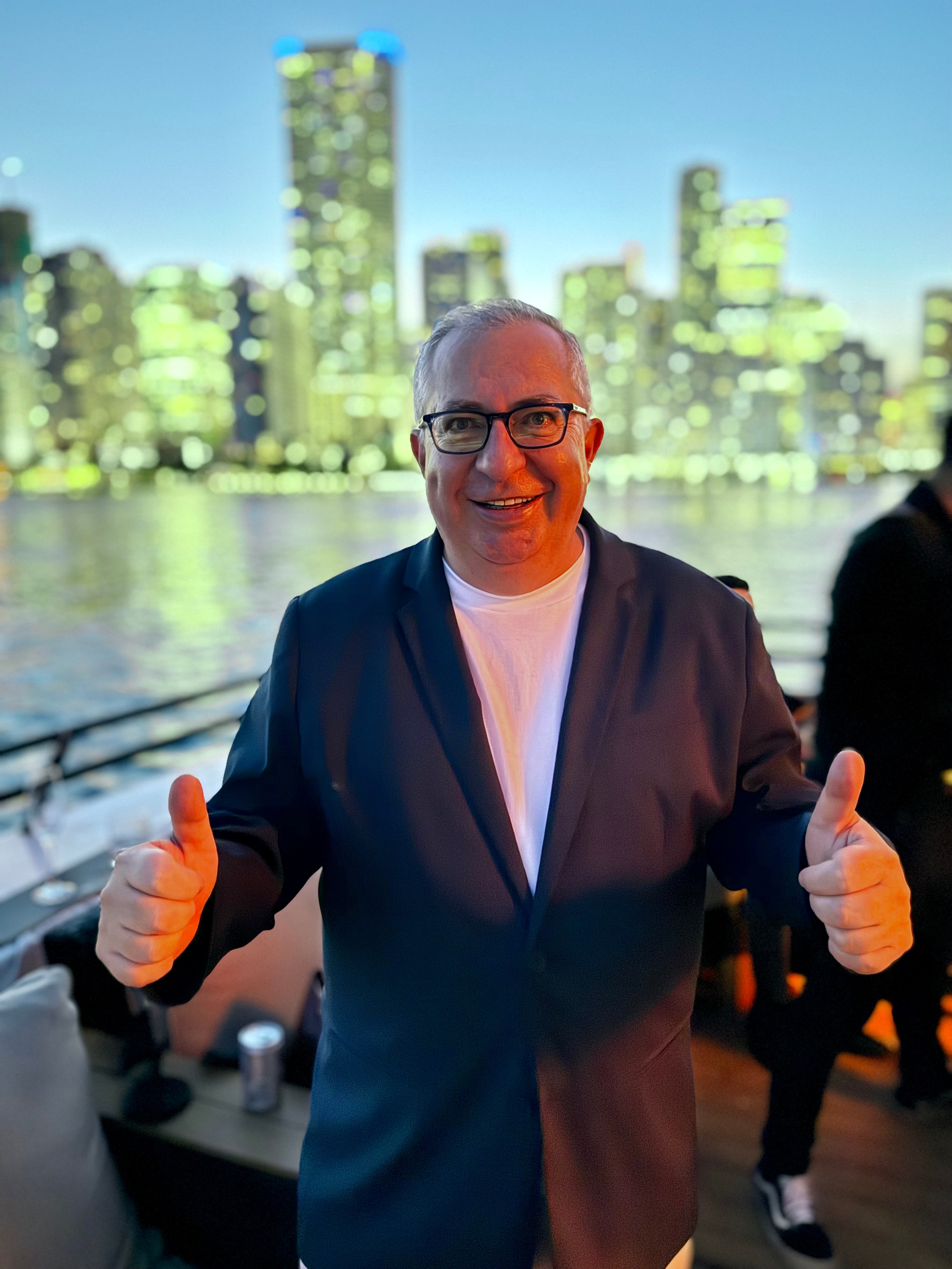Man in glasses and suit smiling with thumbs up, city skyline and water in background at dusk.