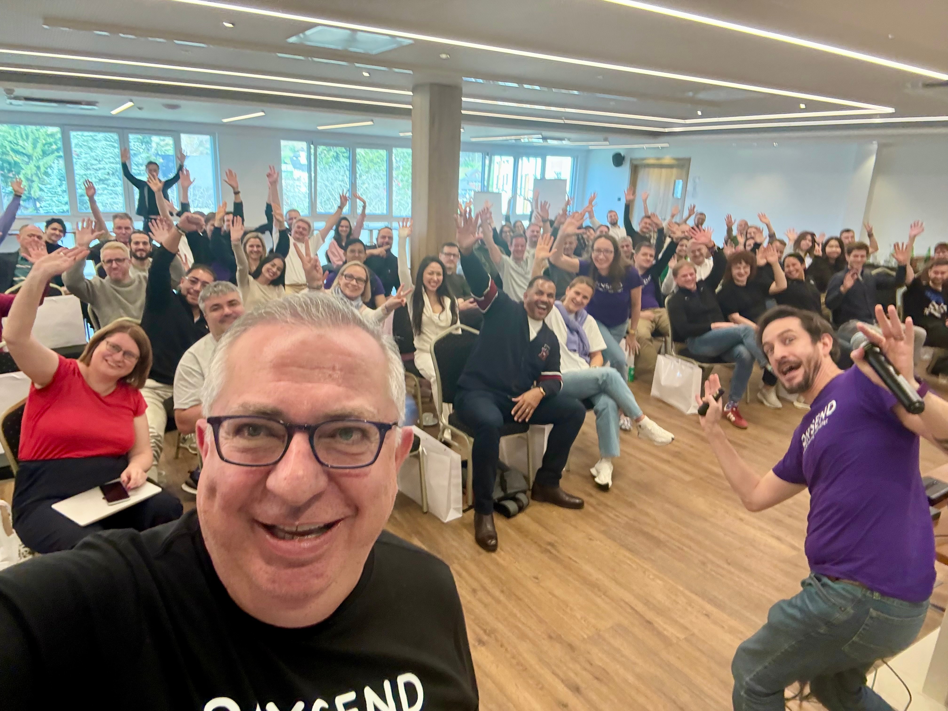 Group of people in a conference room waving at the camera, with a man in the foreground taking a selfie.