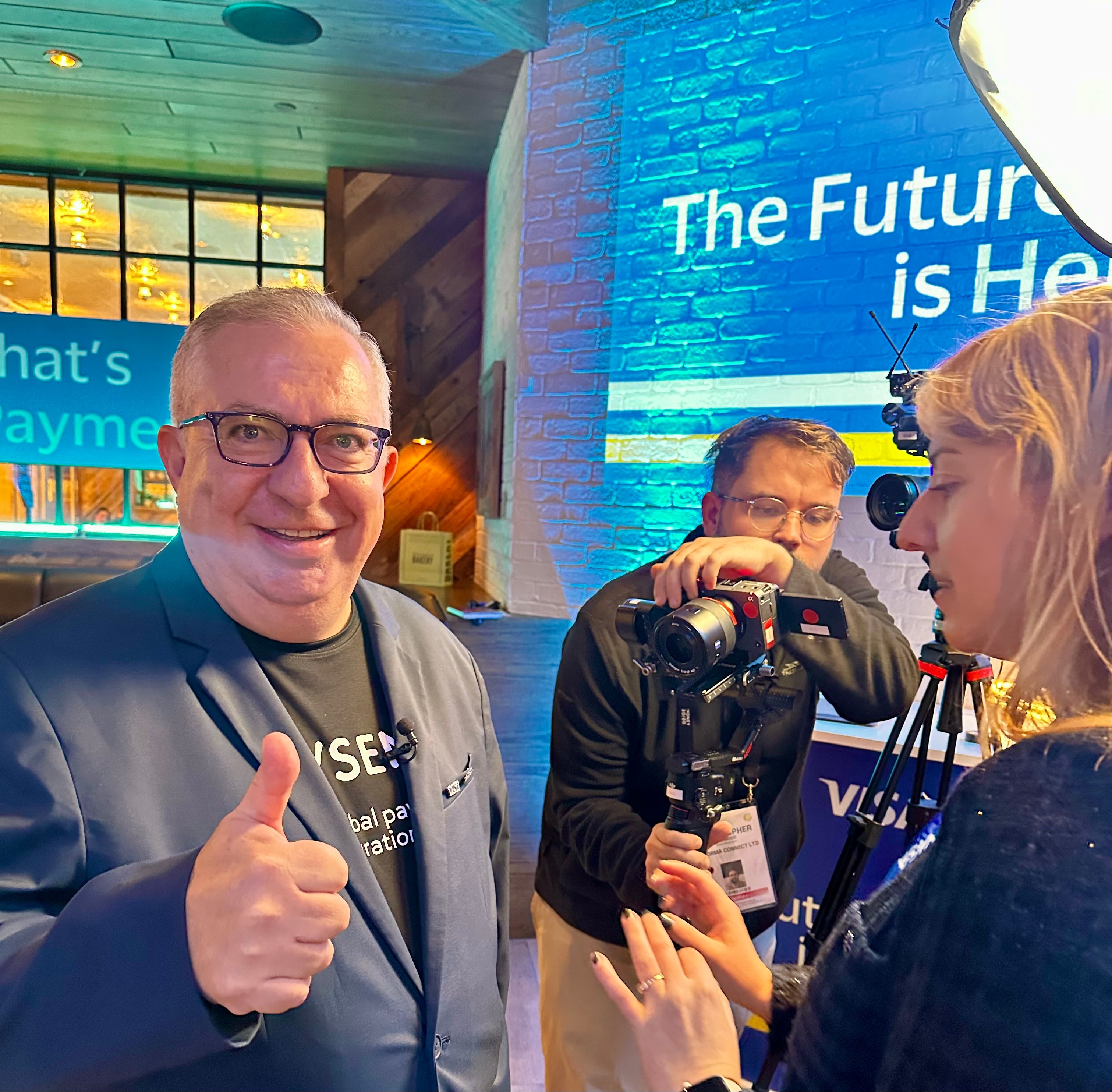 Three people at a technology event, one smiling man giving a thumbs-up, two others with cameras, blue background with text.