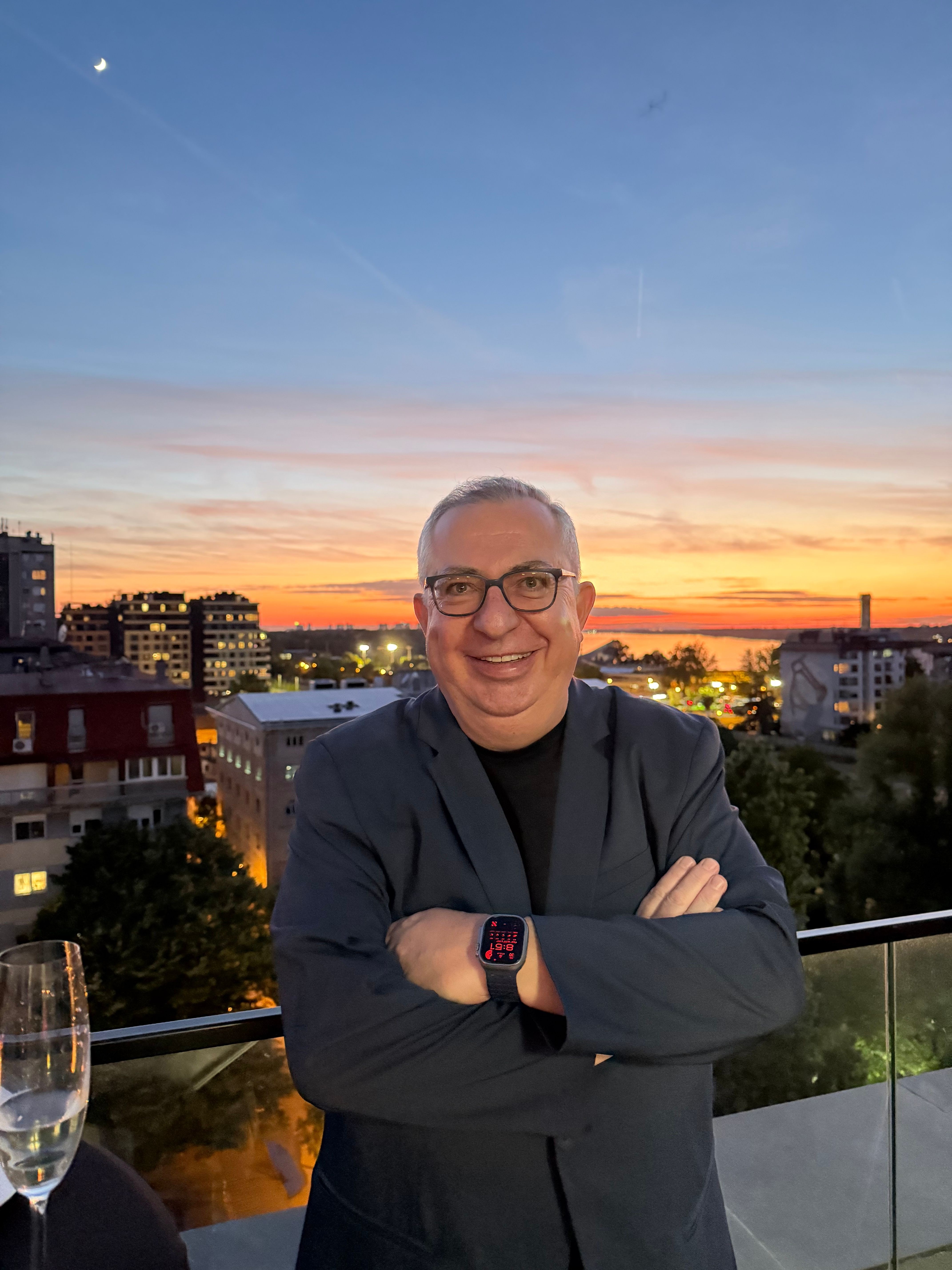 Person with glasses and smartwatch standing on balcony during sunset, city skyline in background.