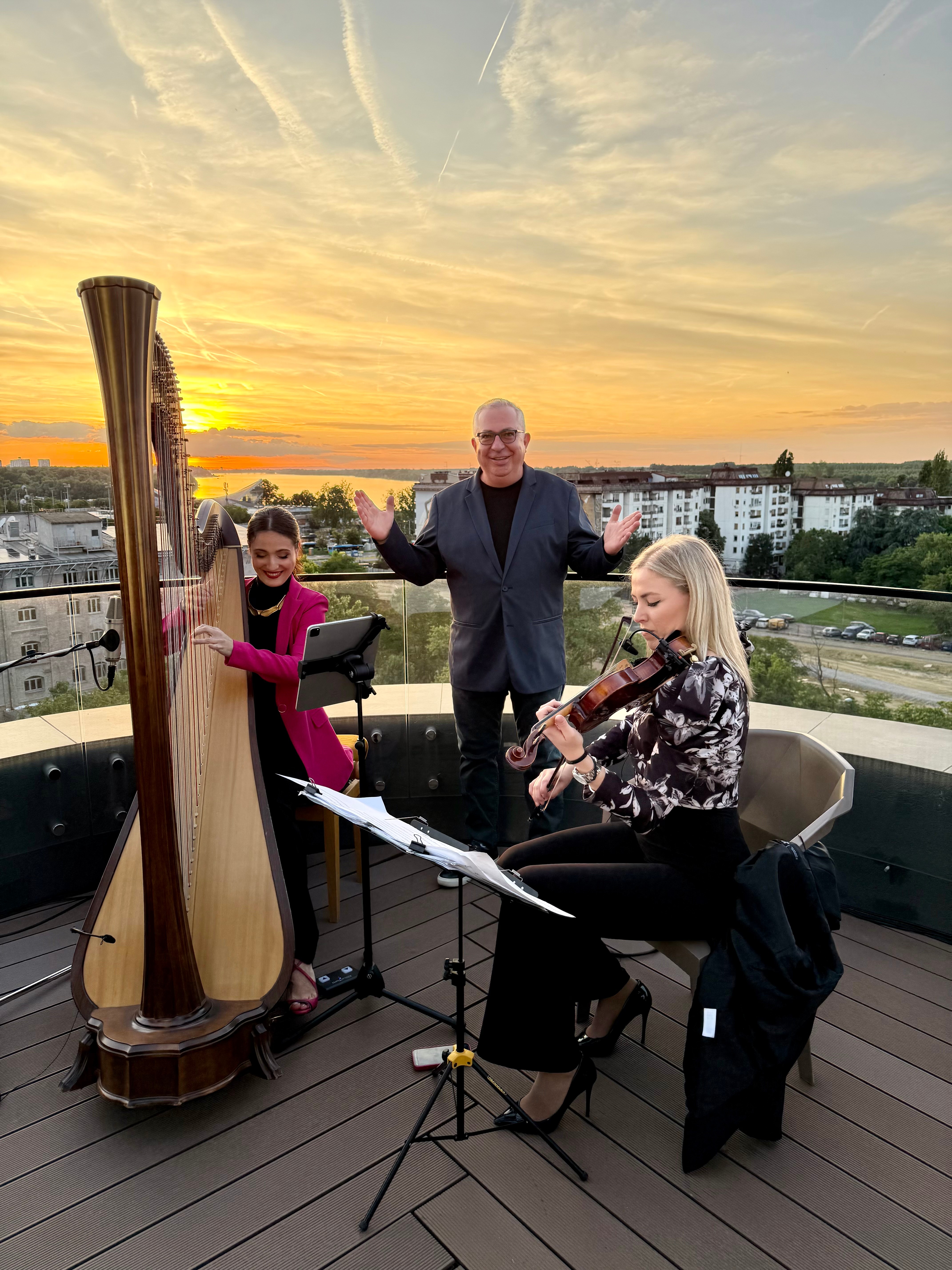 Three people performing music on a rooftop during sunset, with a cityscape in the background.