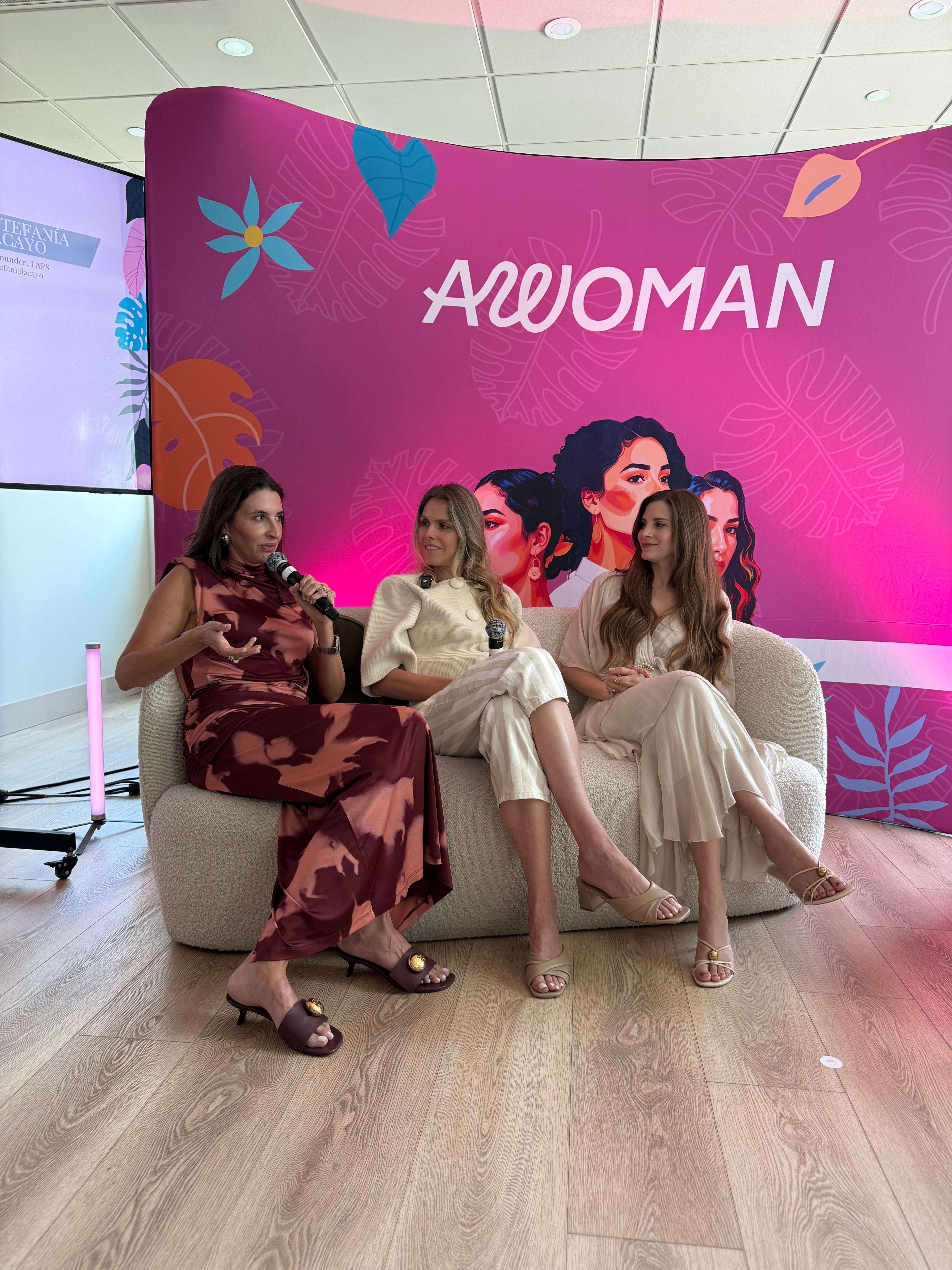 Three women sit on a white sofa in front of a pink backdrop with the word 'AWOMAN' and illustrations of women and flowers.