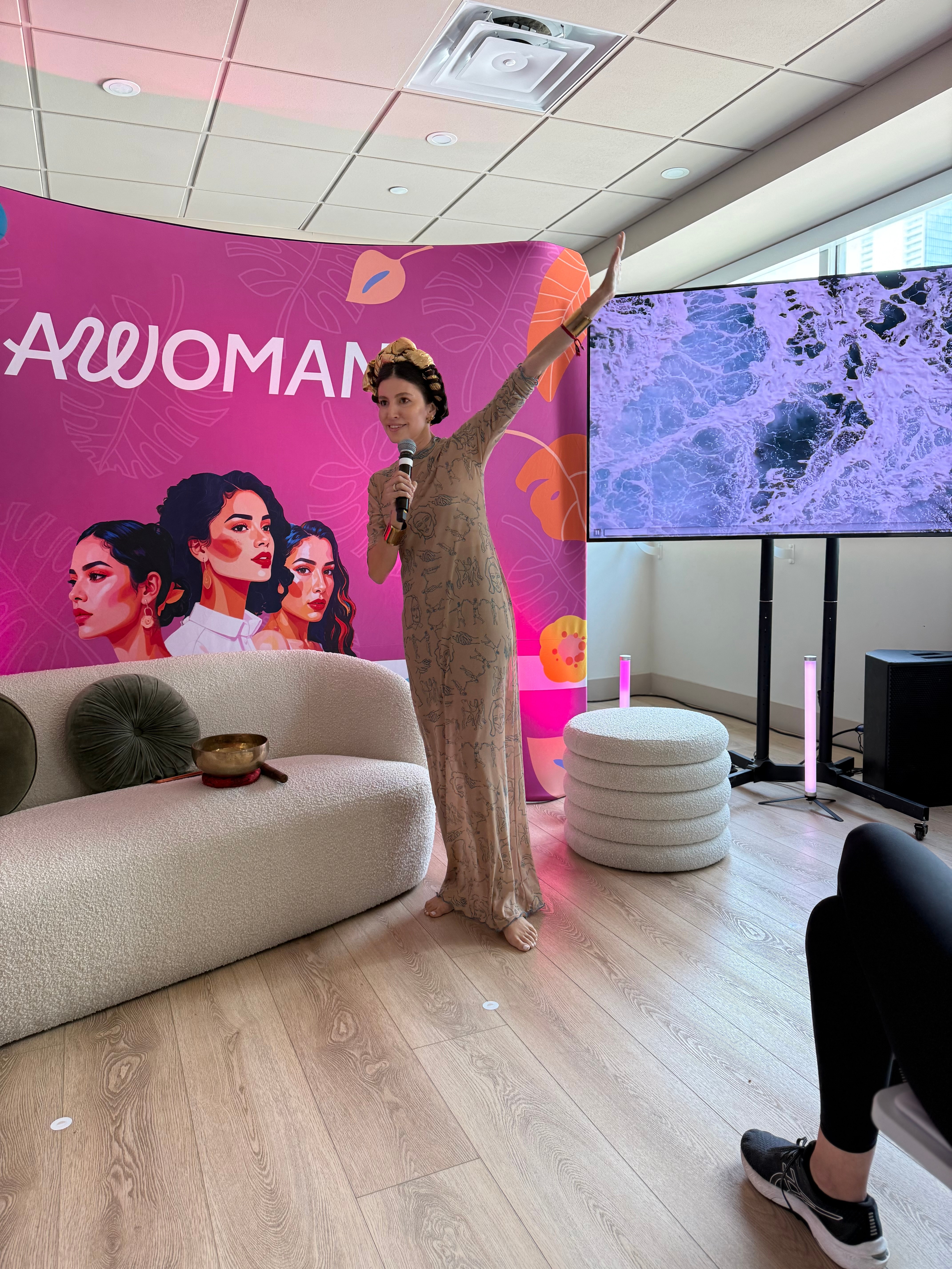 Woman in a long dress standing with one arm raised in front of a pink backdrop with illustrated women and text, next to a large screen.