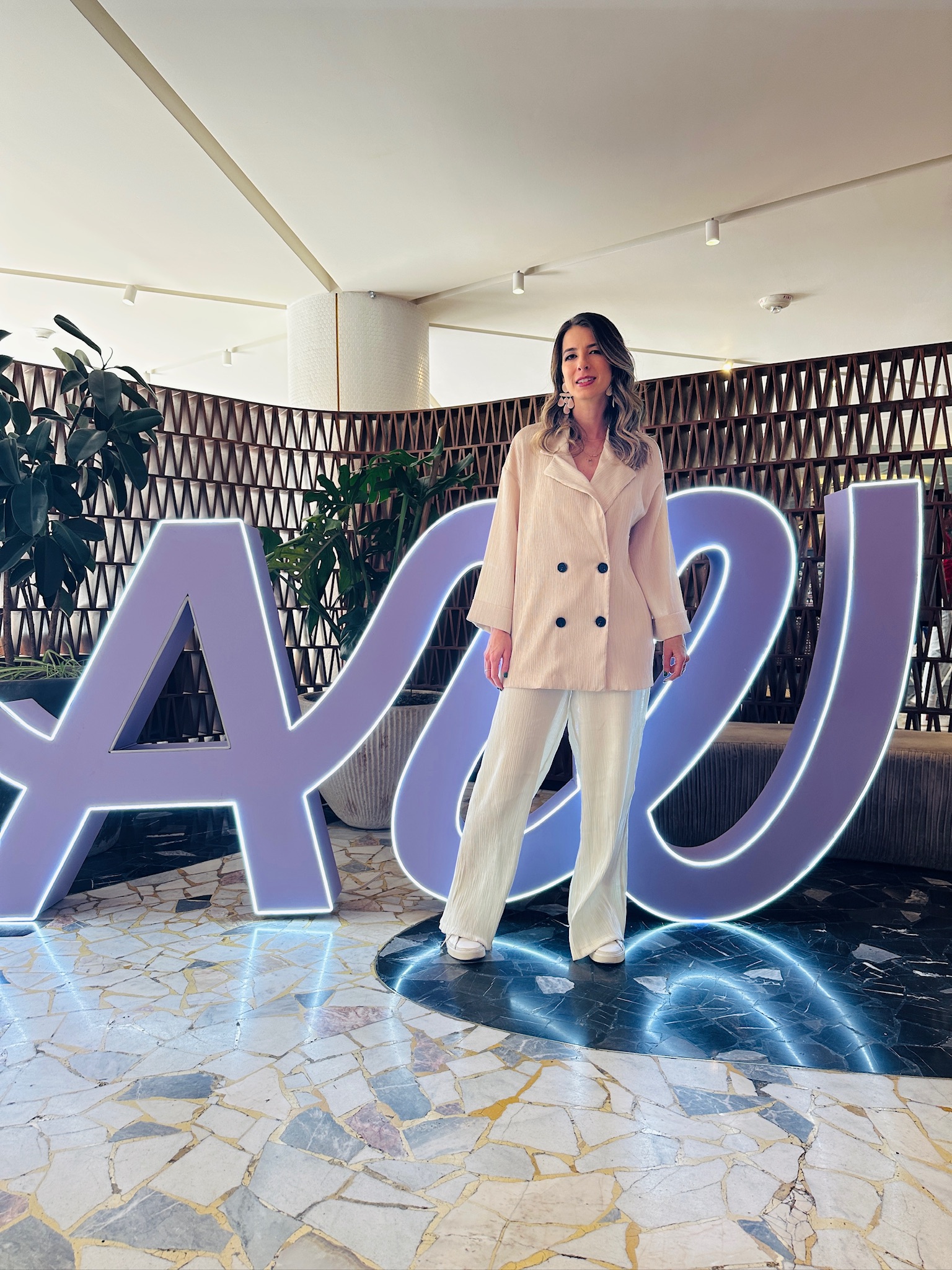 Woman in white suit standing in front of illuminated large letters, indoor setting with plants and patterned wall behind.