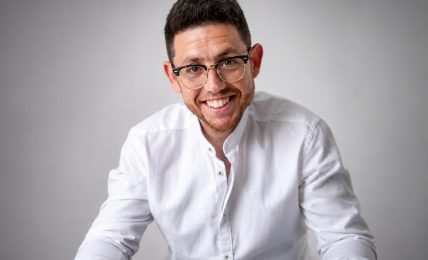 Smiling man with glasses in a white shirt against a plain background.