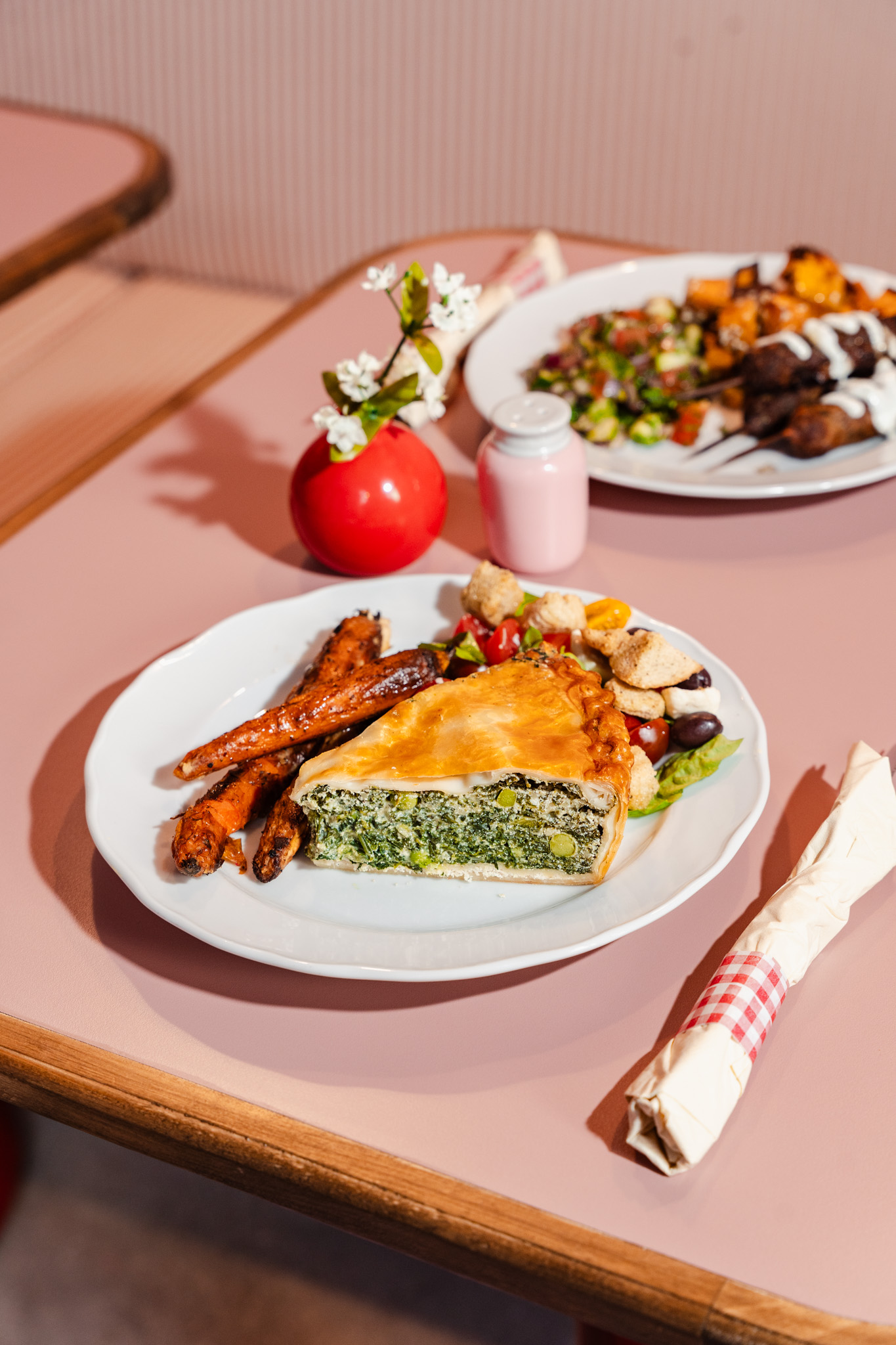 Plate with grilled chicken, vegetables, and a slice of pie on a pink table, with a rolled napkin nearby.