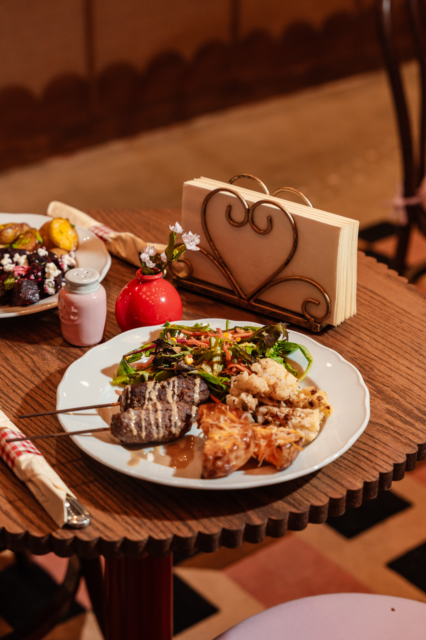 Plate with grilled meat, vegetables, and rice on a wooden table, with a heart-shaped card, flowers, and a small bottle in the background.