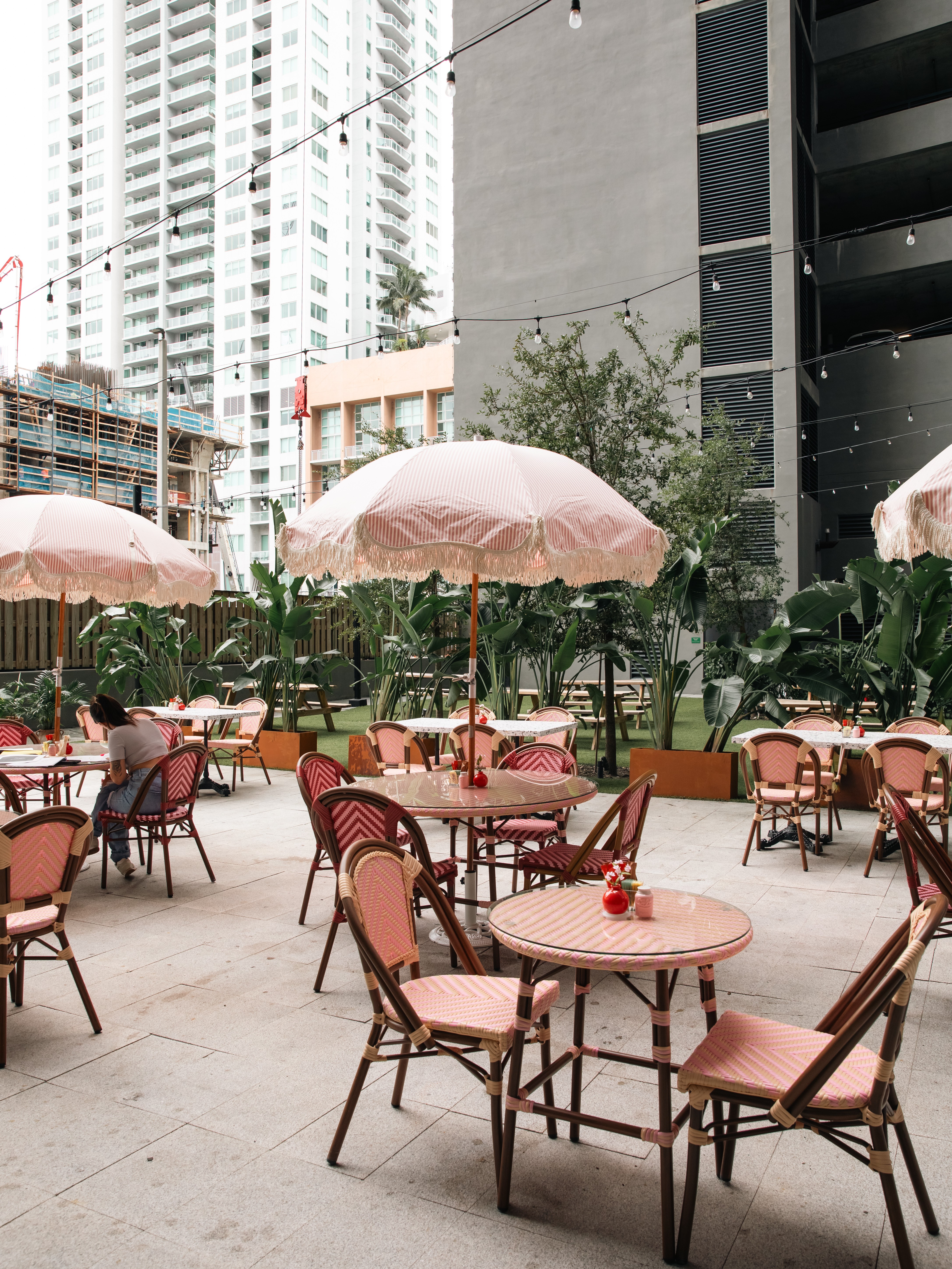 Outdoor seating area with tables, chairs, and pink umbrellas in an urban setting with tall buildings.