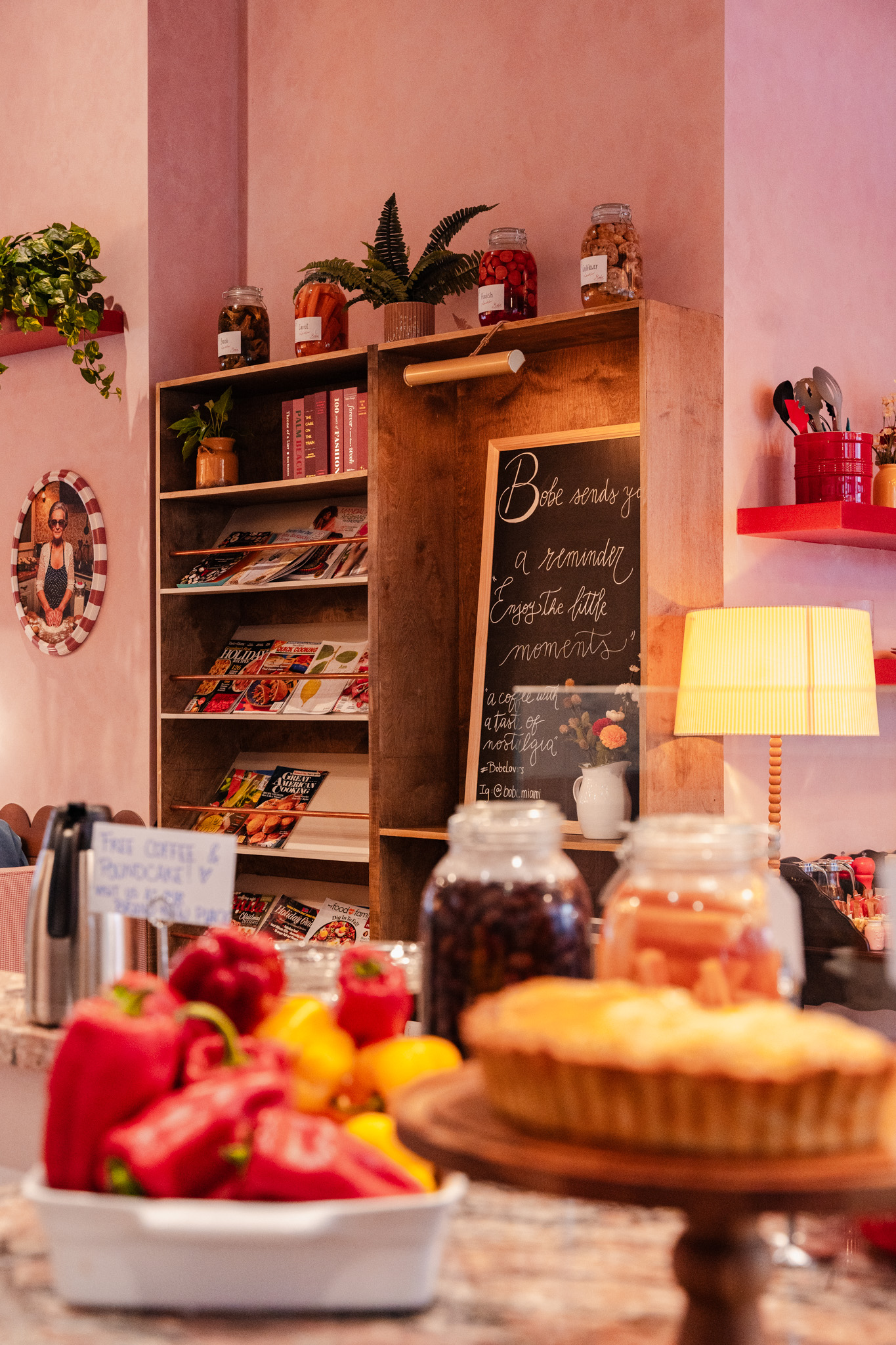 A cozy room with a bookshelf, chalkboard, and table with desserts and fruits, illuminated by a lamp.
