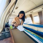 Woman sitting at a desk inside an airplane, working with papers and files, window and overhead compartments visible.