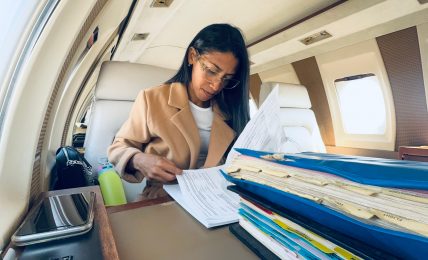 Woman sitting at a desk inside an airplane, working with papers and files, window and overhead compartments visible.