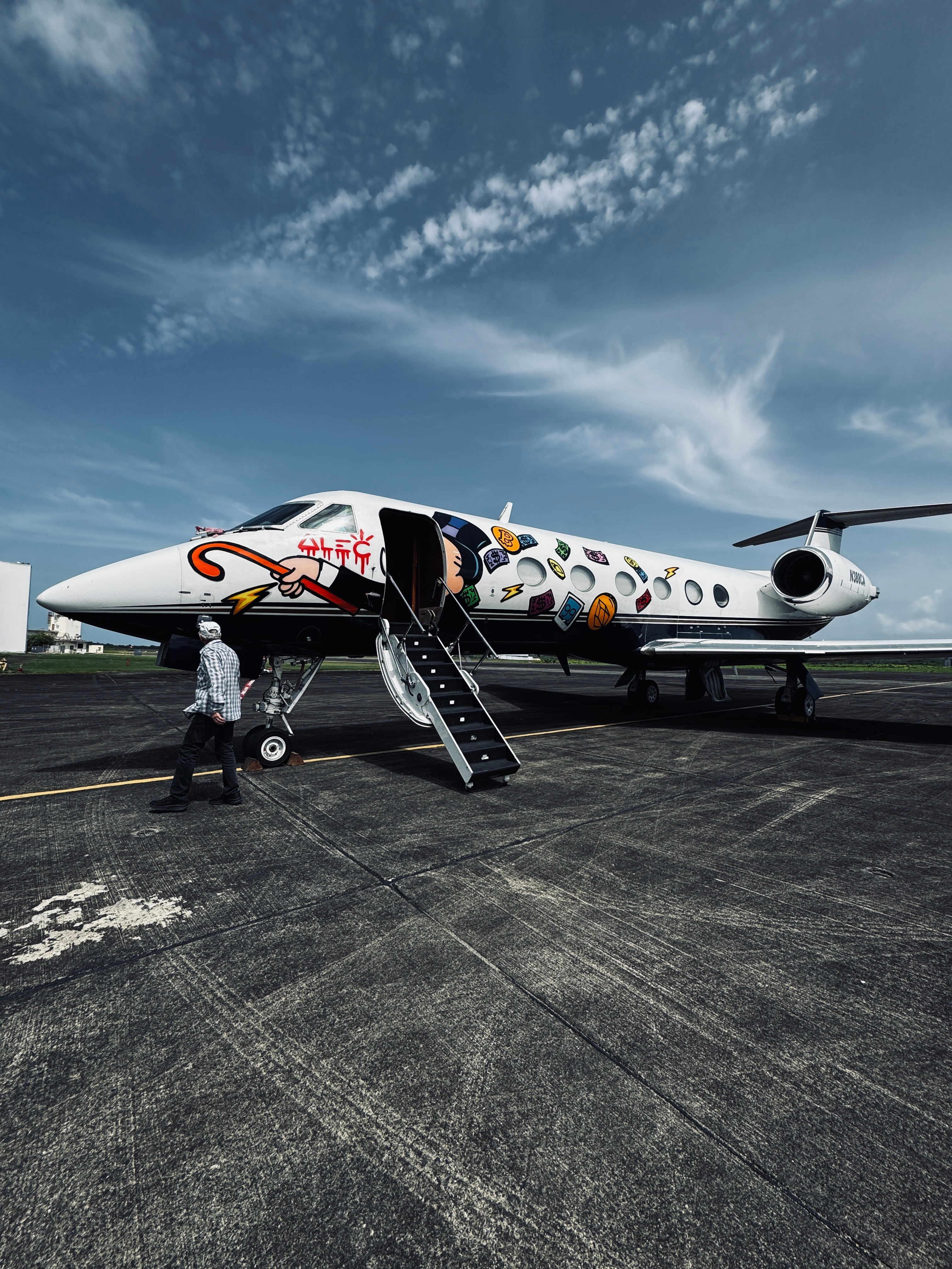 Private jet decorated with colorful patterns on tarmac, with people near the aircraft and stairs, under a partly cloudy sky.