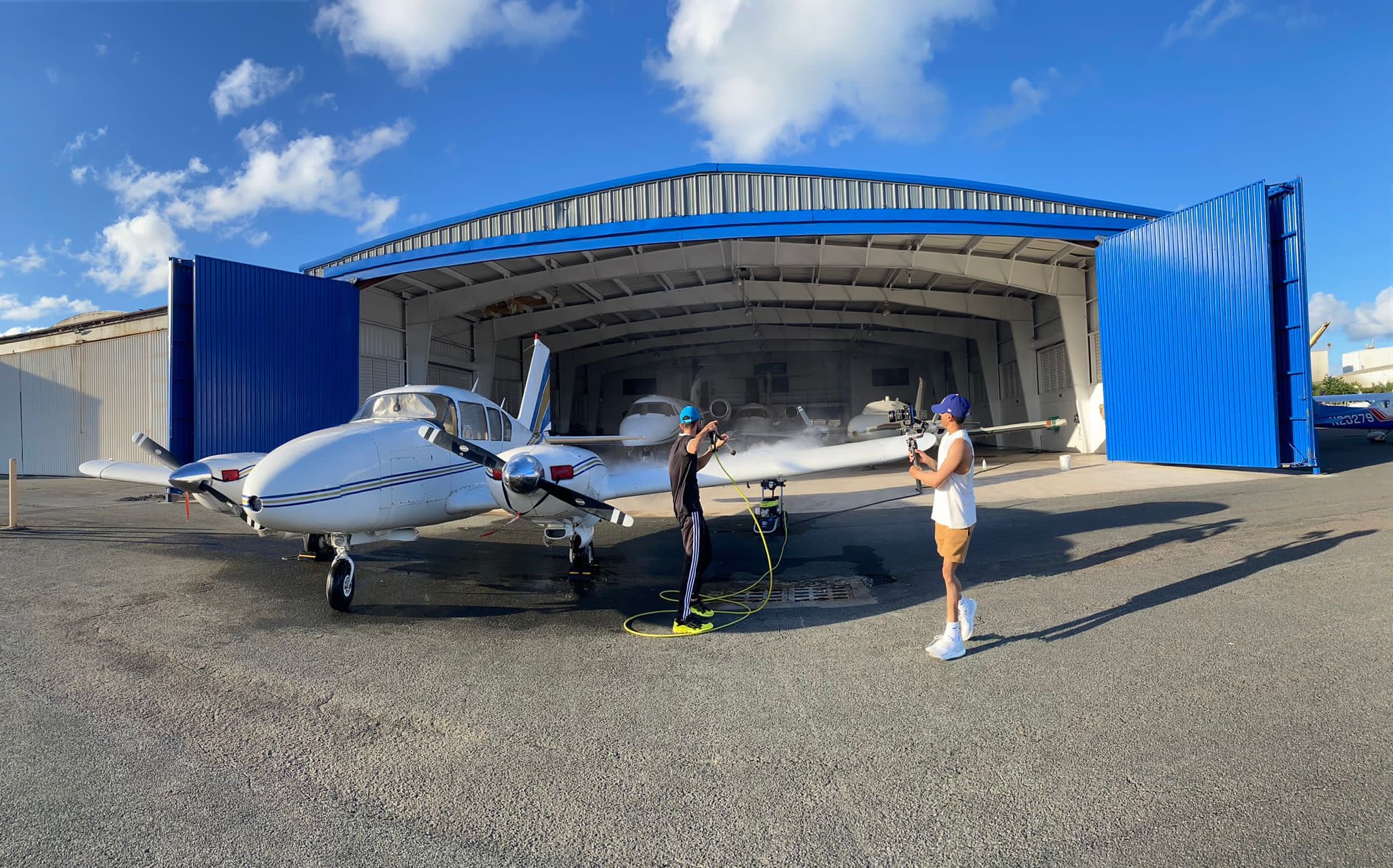 Two people cleaning a small airplane outside a blue hangar on a sunny day.