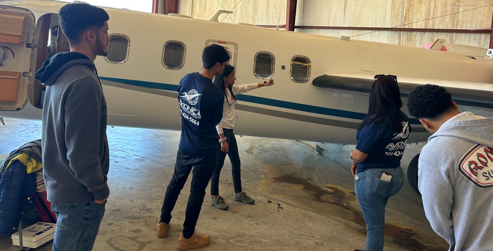 Group of people standing near an airplane inside a hangar, some taking photos or looking at the aircraft.