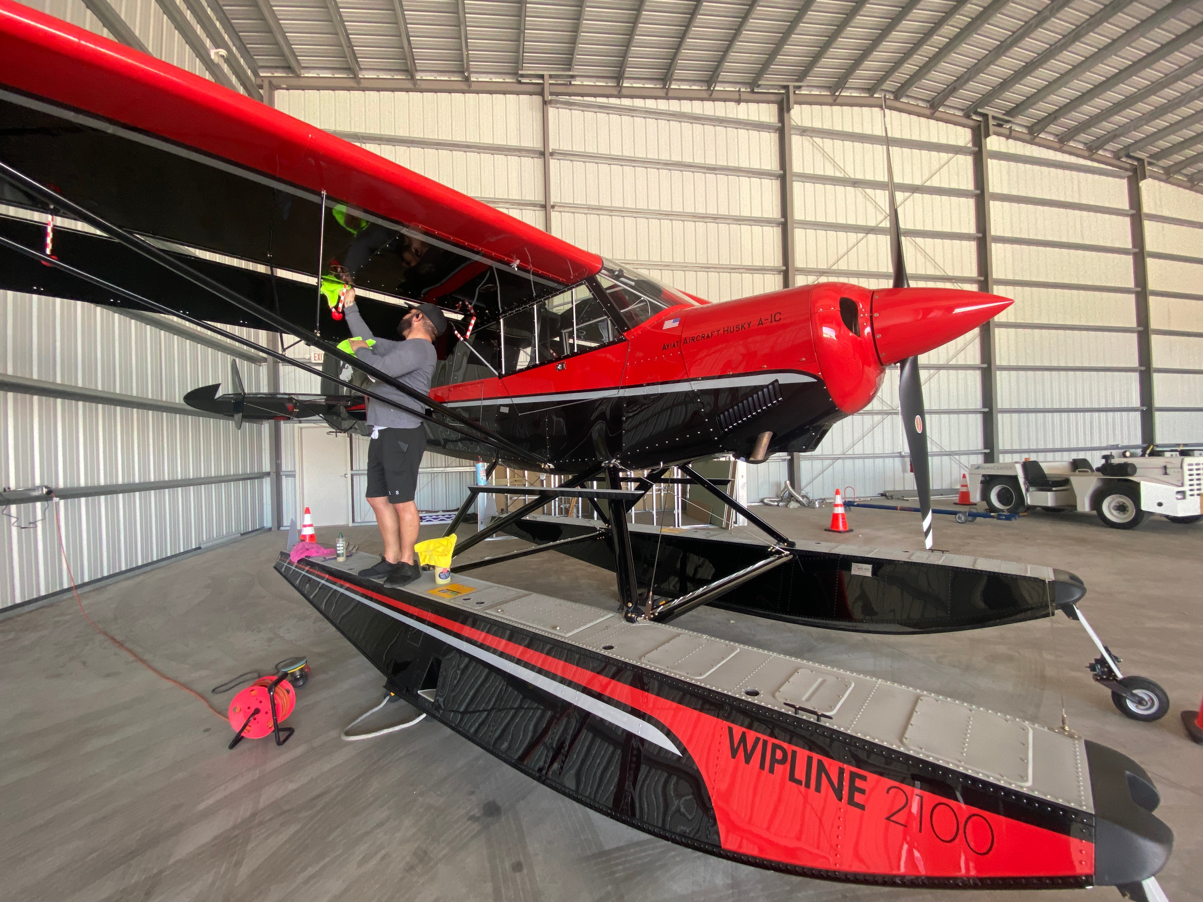 Person working on a red and black aircraft inside a hangar, with a WIPLINE 4100 boat nearby.