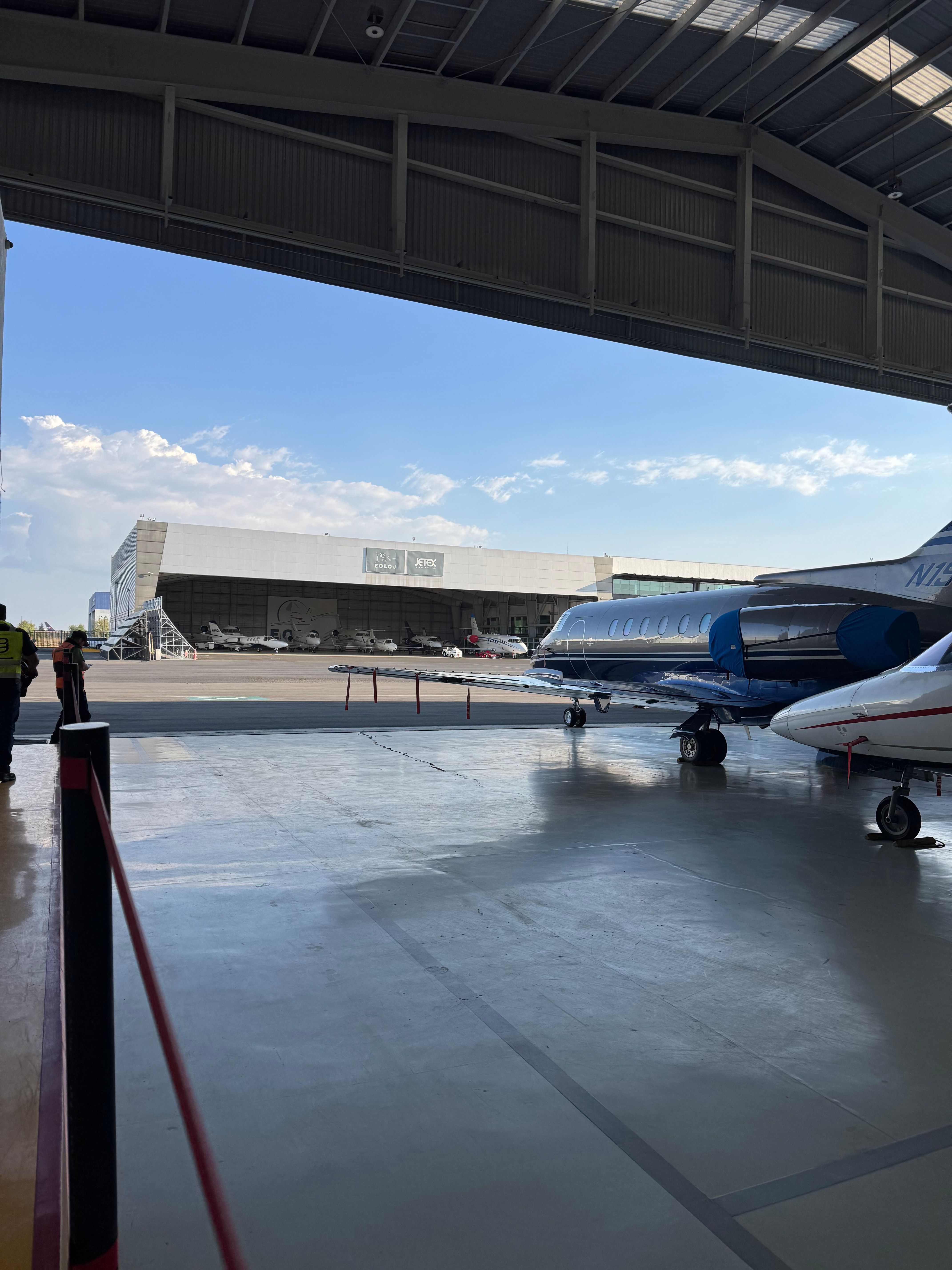 View of a parking area with cars parked, seen from a covered structure, with a blue sky and clouds in the background.