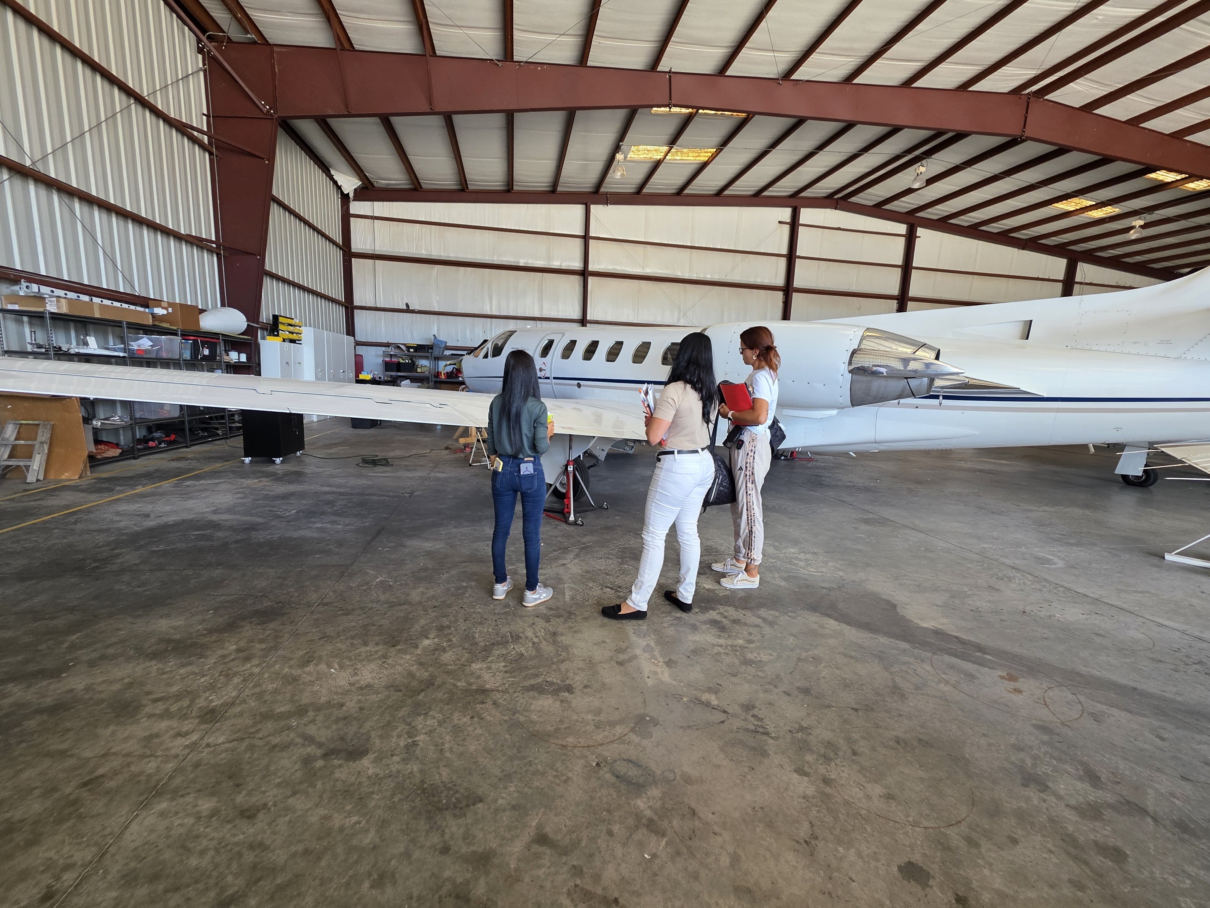 Four people standing near a white private jet inside a hangar, with a curved ceiling and metal walls.