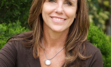 Smiling woman with brown hair outdoors, surrounded by greenery, wearing a dark top and a necklace.