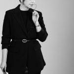 Woman in black blazer and skirt holding a book, looking down thoughtfully, against a plain background.