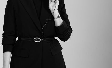Woman in black blazer and skirt holding a book, looking down thoughtfully, against a plain background.