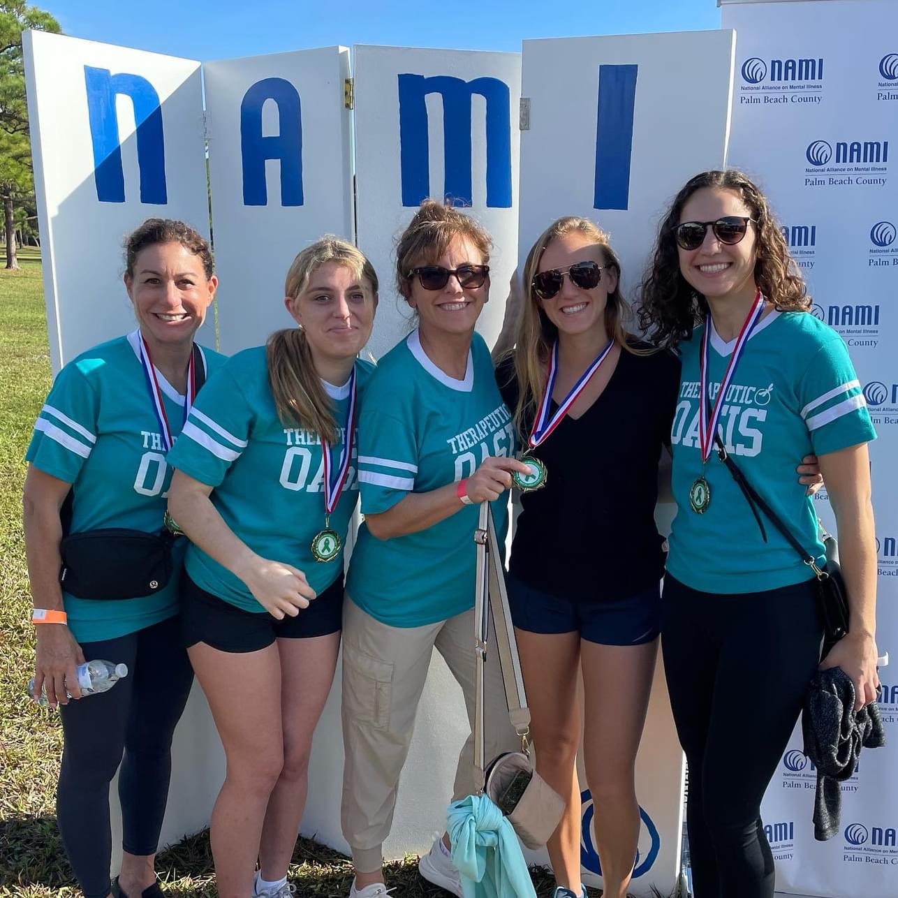Five women in teal sports jerseys with medals, standing outdoors in front of a blue and white backdrop with 'NAMI' letters.
