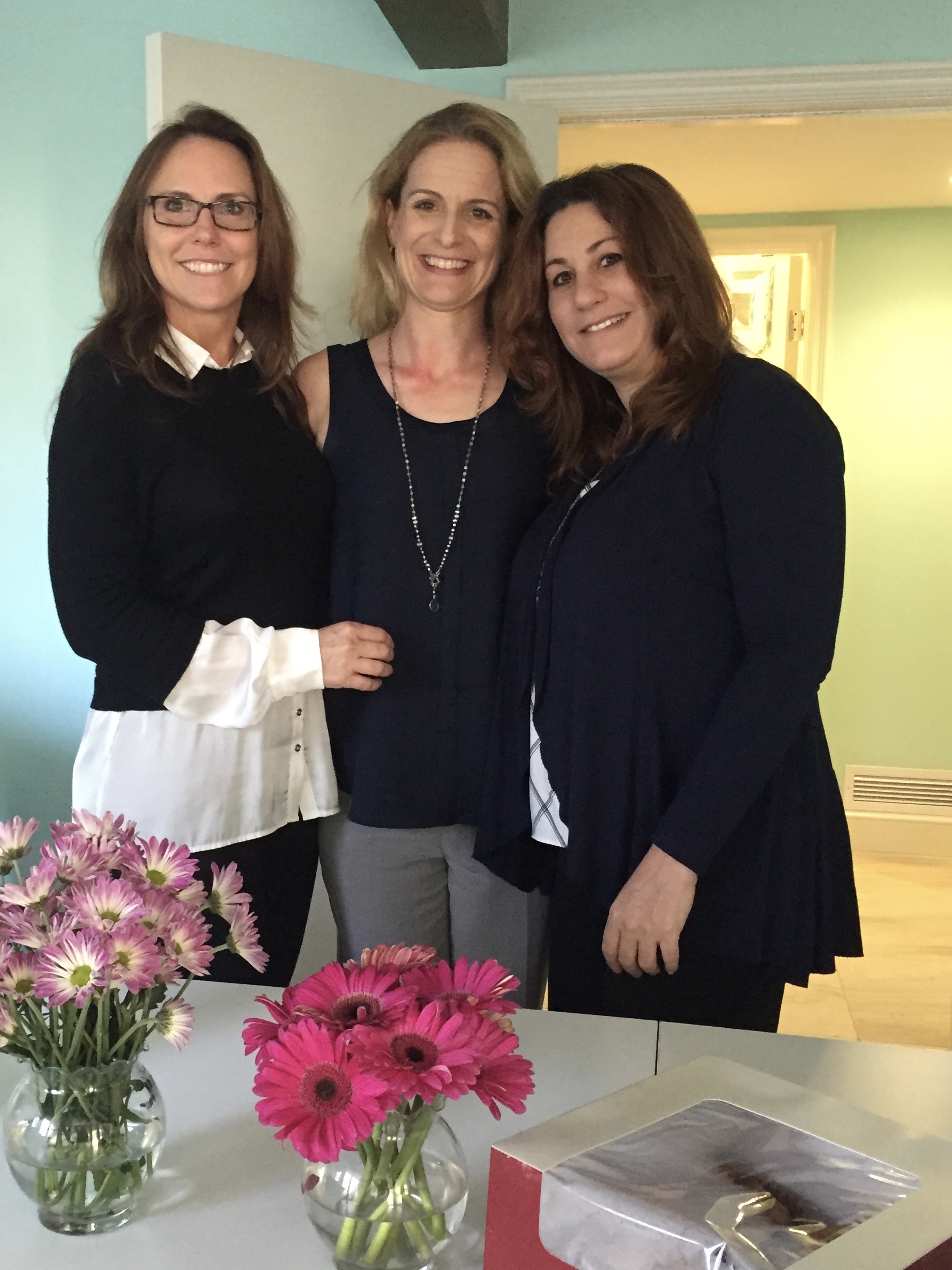 Three women standing indoors, smiling, with pink flowers in vases on a table in front of them.