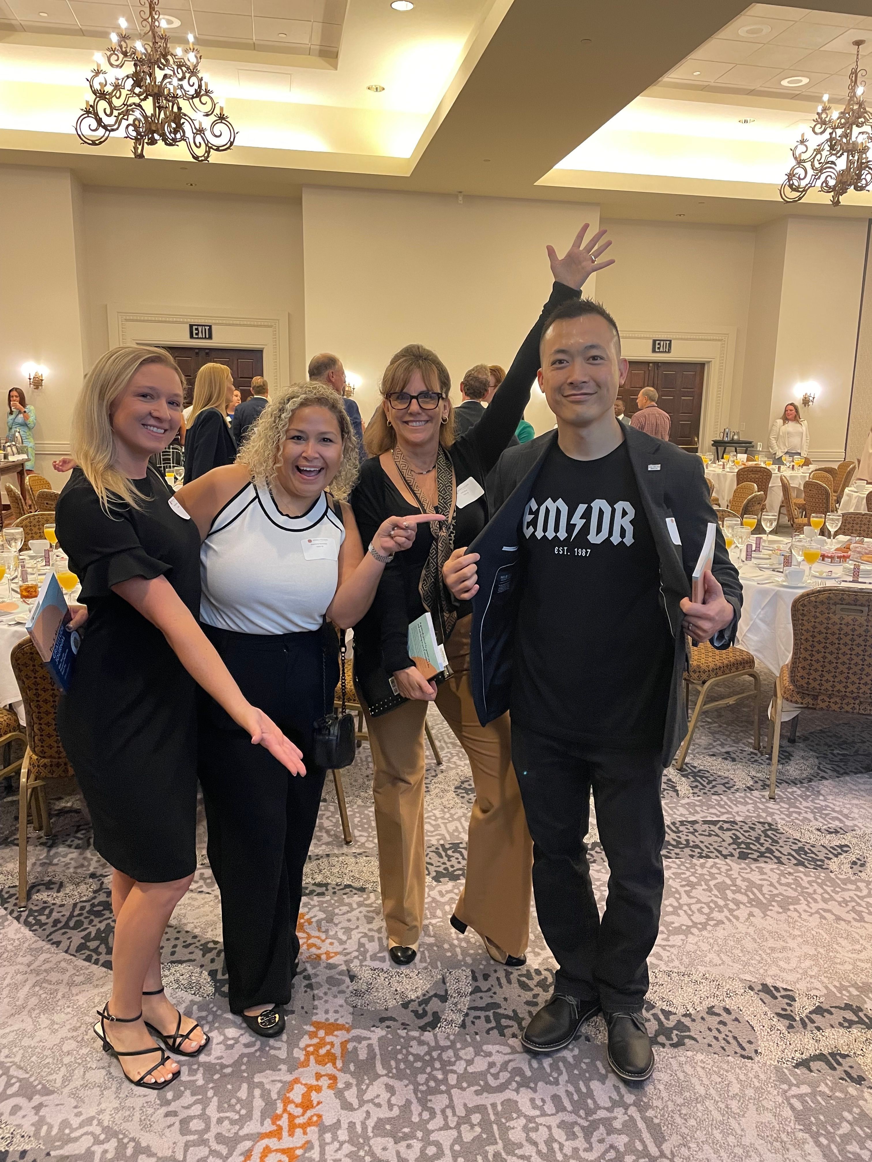 Four people posing together at an indoor event, smiling and making gestures, with tables and decorations in the background.