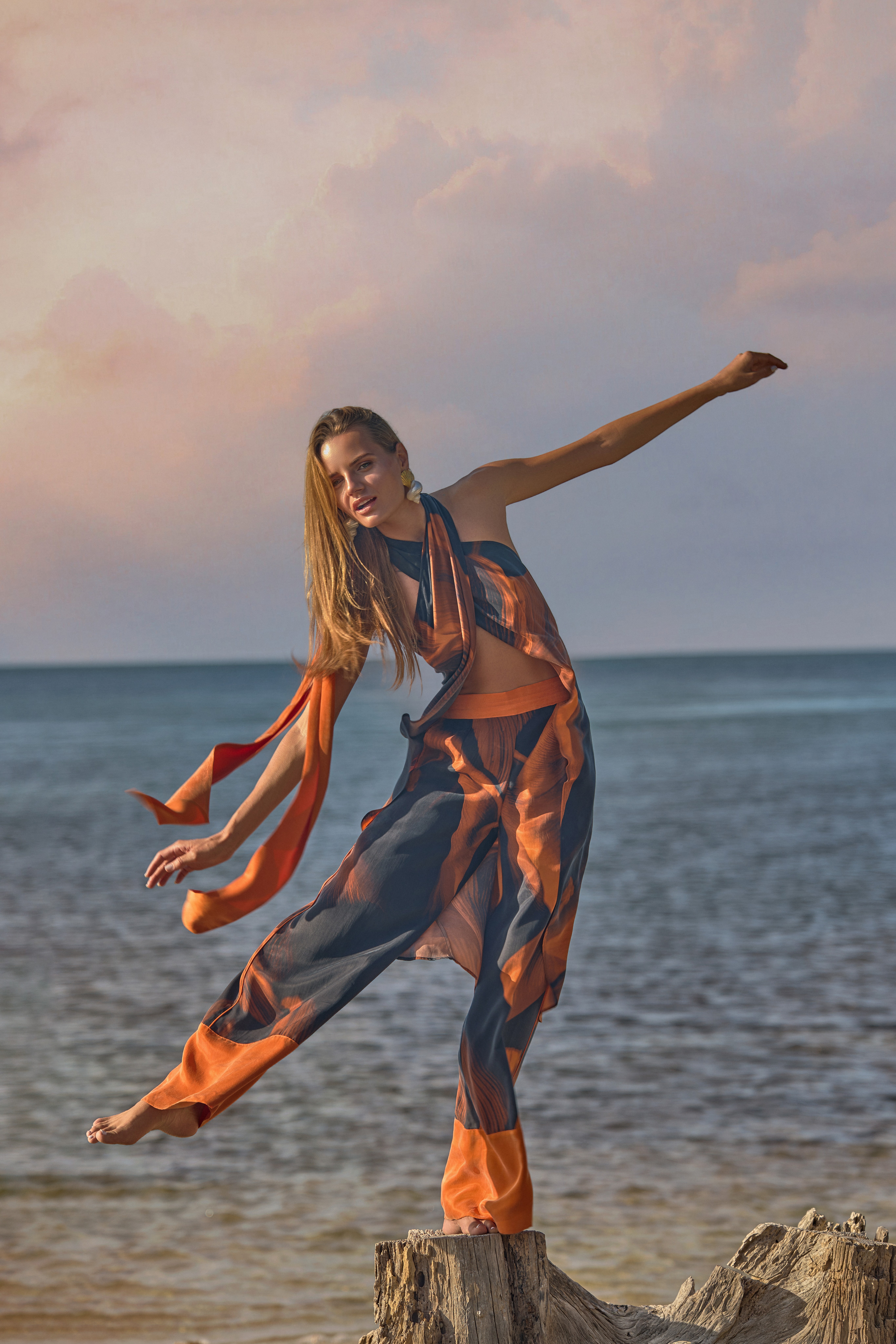 Person balancing on a tree stump at the beach during sunset, arms extended, wearing colorful loose clothing.