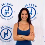 Woman with dark hair smiling, arms crossed, standing in front of Jetset Modern Pilates logo backdrop.