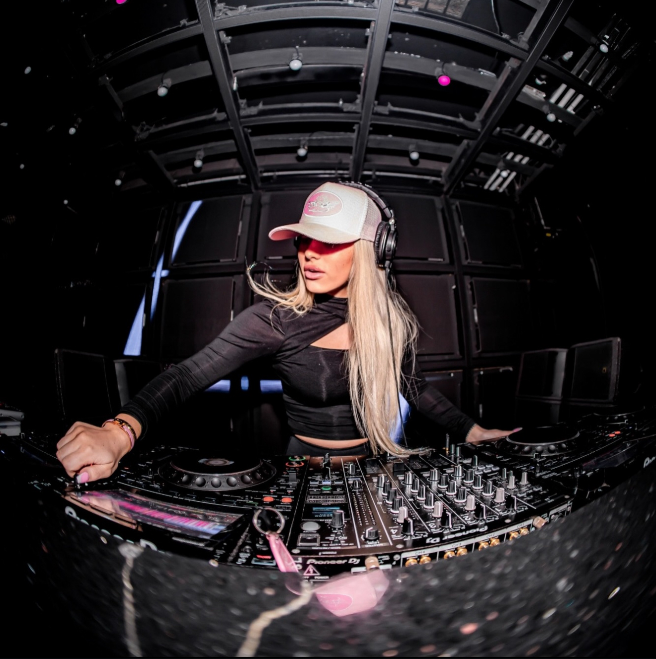 Female DJ wearing headphones and a cap, operating DJ equipment in a dark club setting.