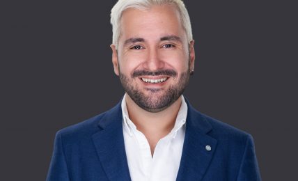 Portrait of a smiling man with white hair and beard wearing a blue suit and white shirt against a dark background.