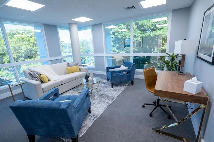 Modern office with large windows, white sofa, blue armchairs, wooden desk, and potted plant, bright and airy space.