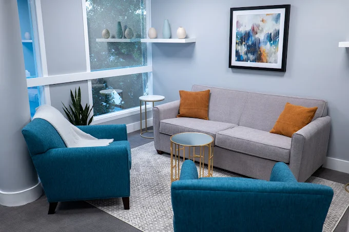 Living room with a gray sofa, two blue armchairs, a small round table, and wall art, with a window and shelves.