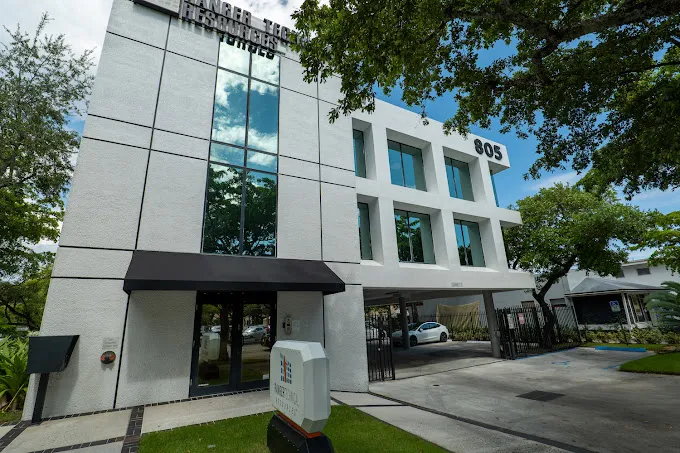 Modern building with large windows and trees in front, number 805 visible, under a partly cloudy sky.