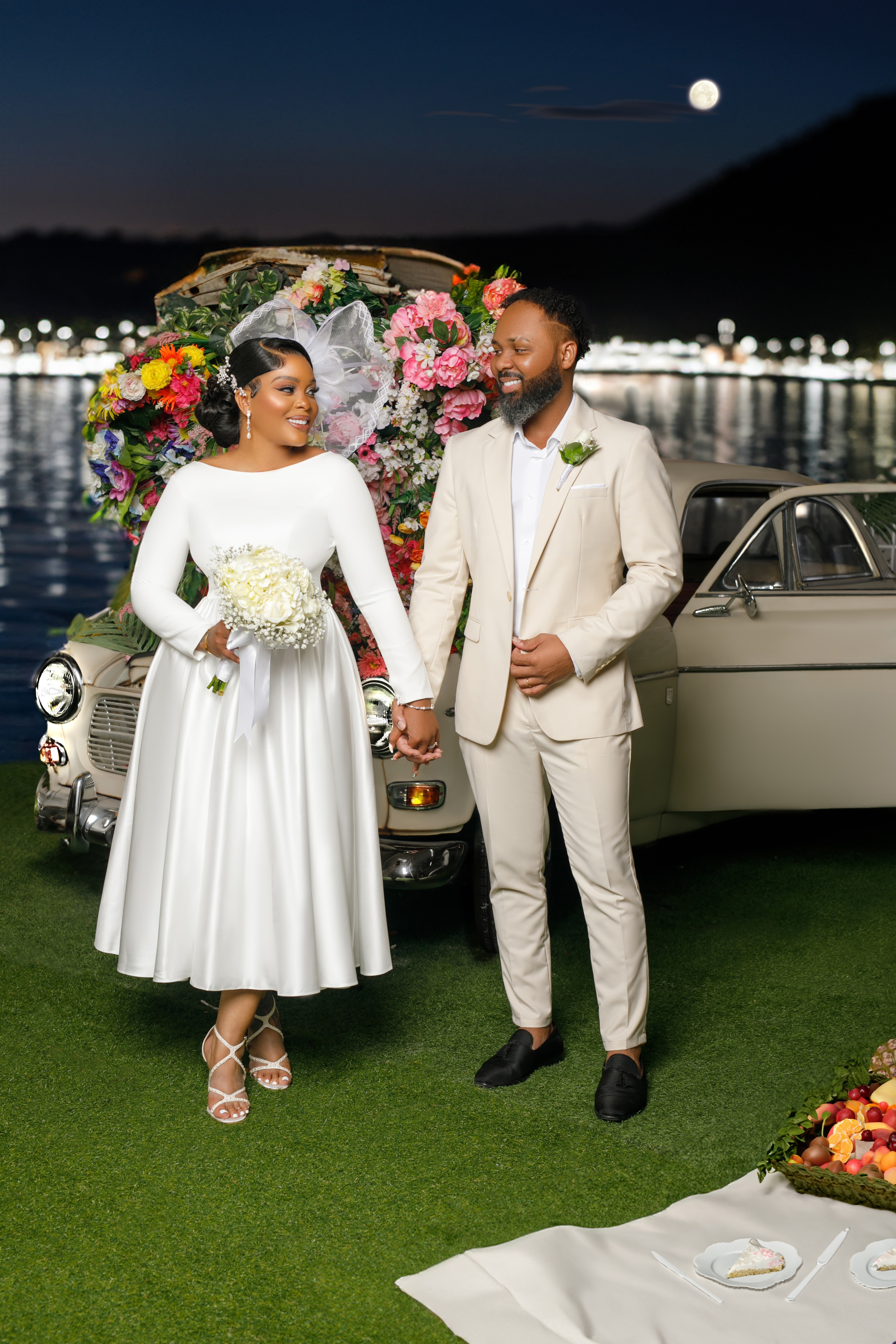 A couple in wedding attire holding hands outdoors at night with a decorated car and a full moon in the sky.
