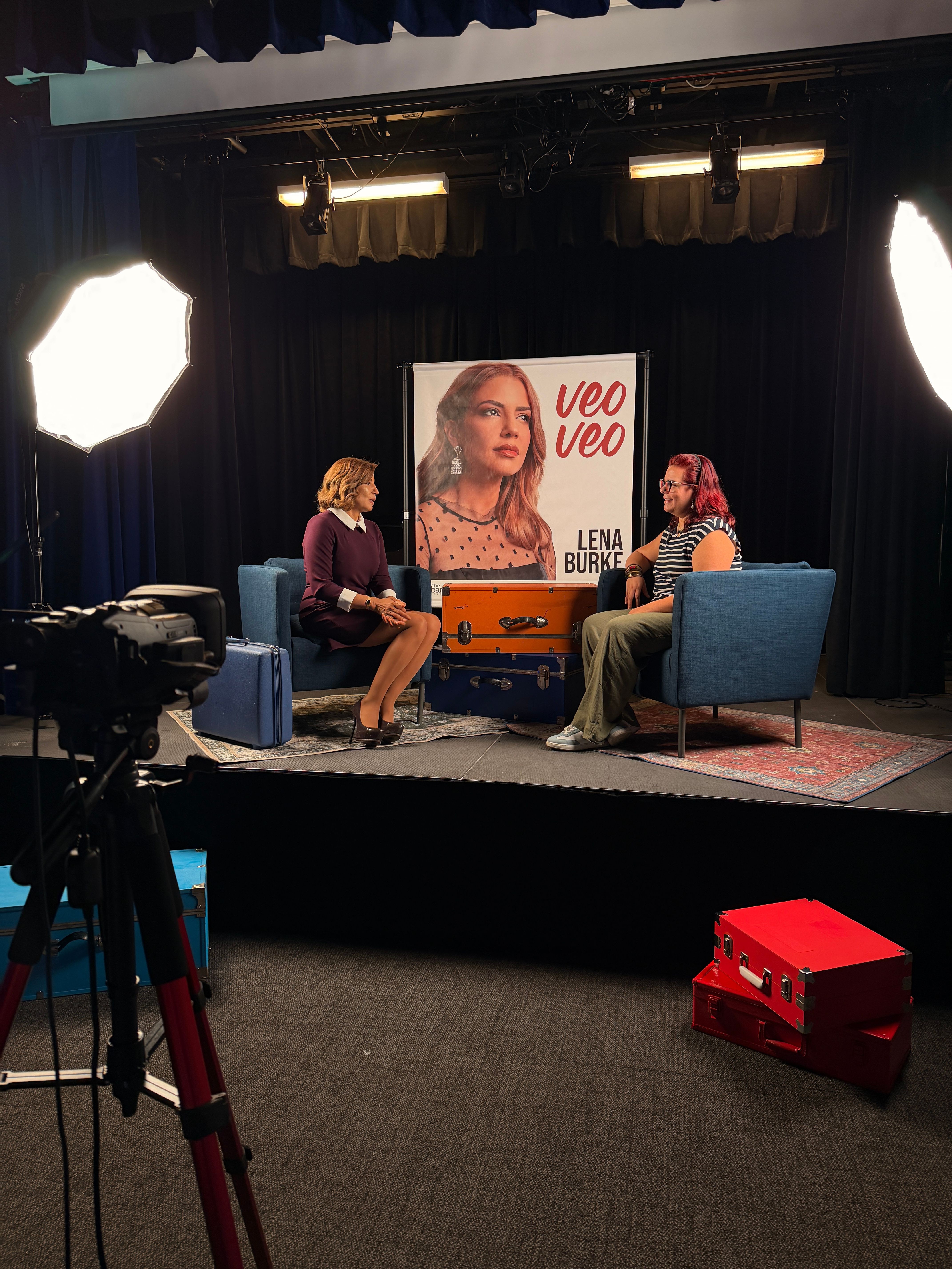 Two women sit on chairs on a stage with a large poster behind them, one woman holds a microphone, stage lighting visible.