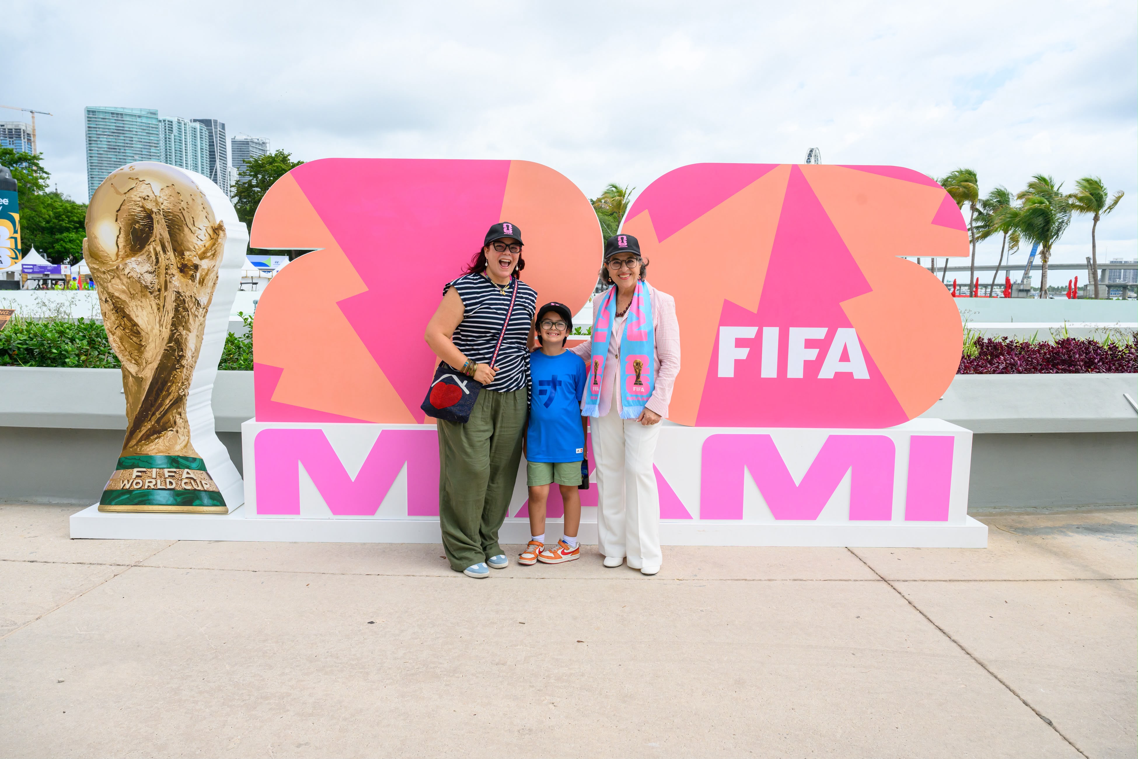 Three people stand in front of a pink and orange FIFA World Cup sign with a trophy statue on the left, outdoors with palm trees and buildings in the background.