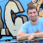 Young man with short hair and a beard smiling, wearing a light blue shirt, leaning on a yellow railing in front of graffiti art.