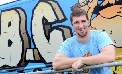Young man with short hair and a beard smiling, wearing a light blue shirt, leaning on a yellow railing in front of graffiti art.