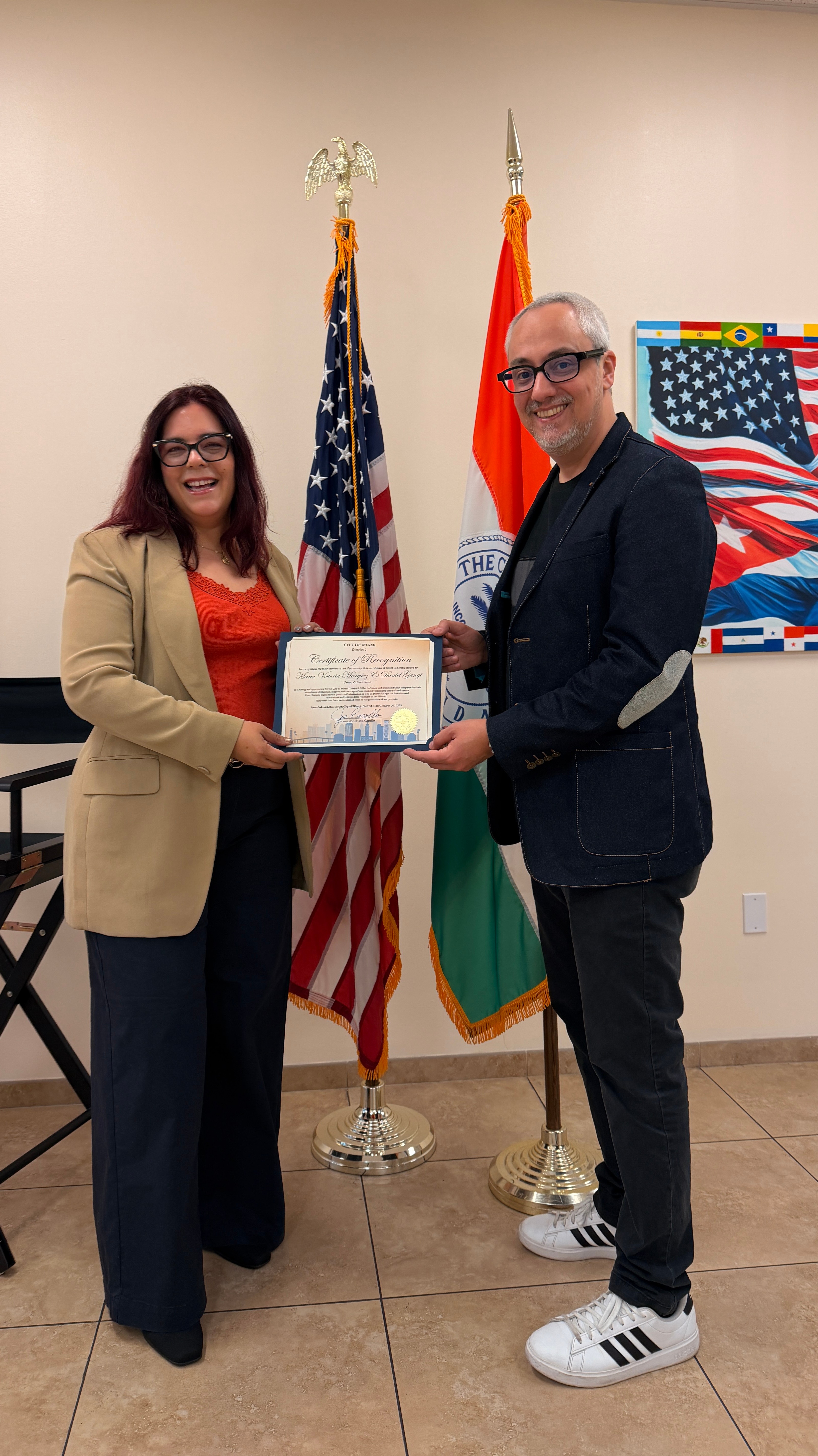 Two people stand indoors holding a certificate, with American and another flag behind them, and a colorful artwork on the wall.