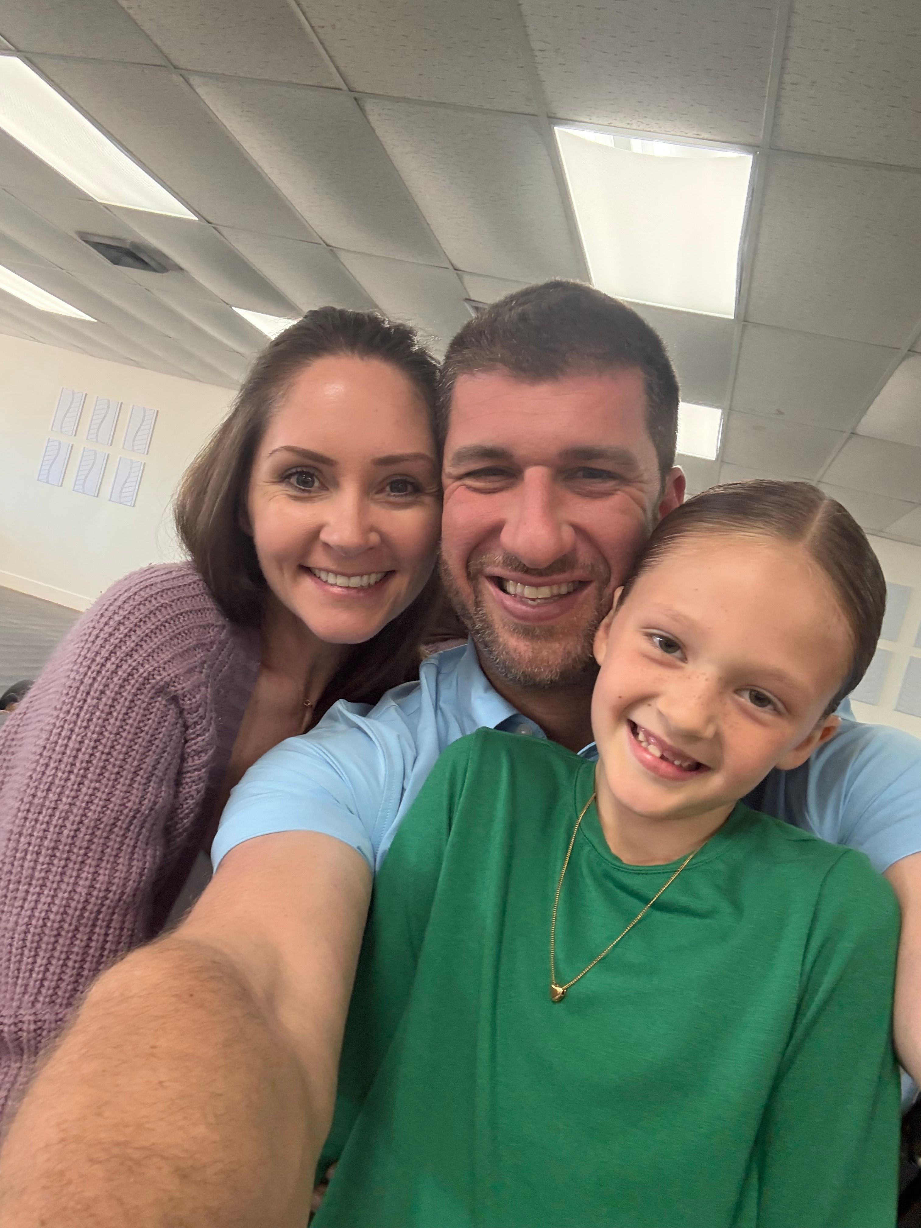 A smiling man, woman, and child taking a selfie together indoors, ceiling lights visible in the background.